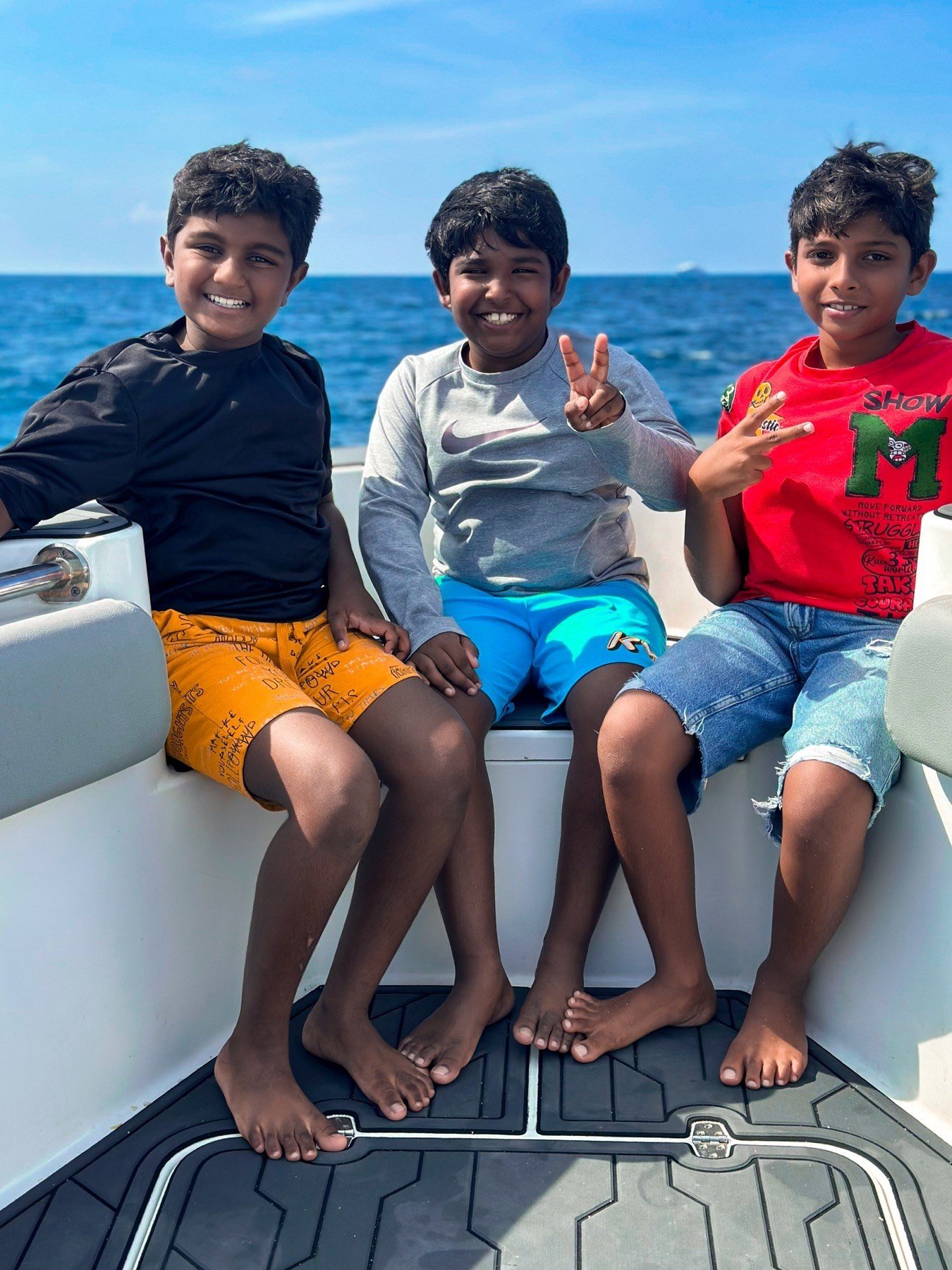 Three boys sitting on a boat with the ocean in the background, smiling and making peace signs.