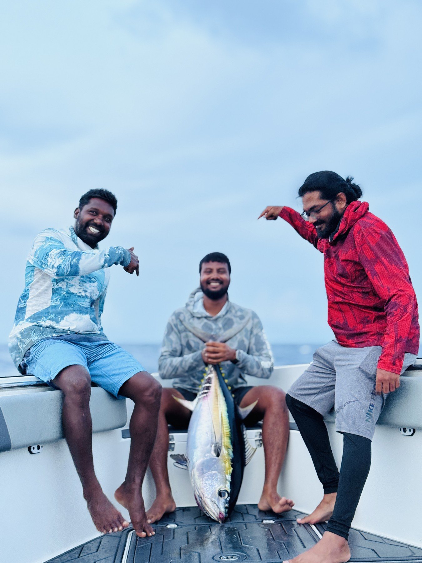 Three happy men on a boat with a large fish. One man is sitting with his arm around the fish, another standing on the right, and a third sitting in the middle. They are smiling and pointing at the fish, against a cloudy sky background.