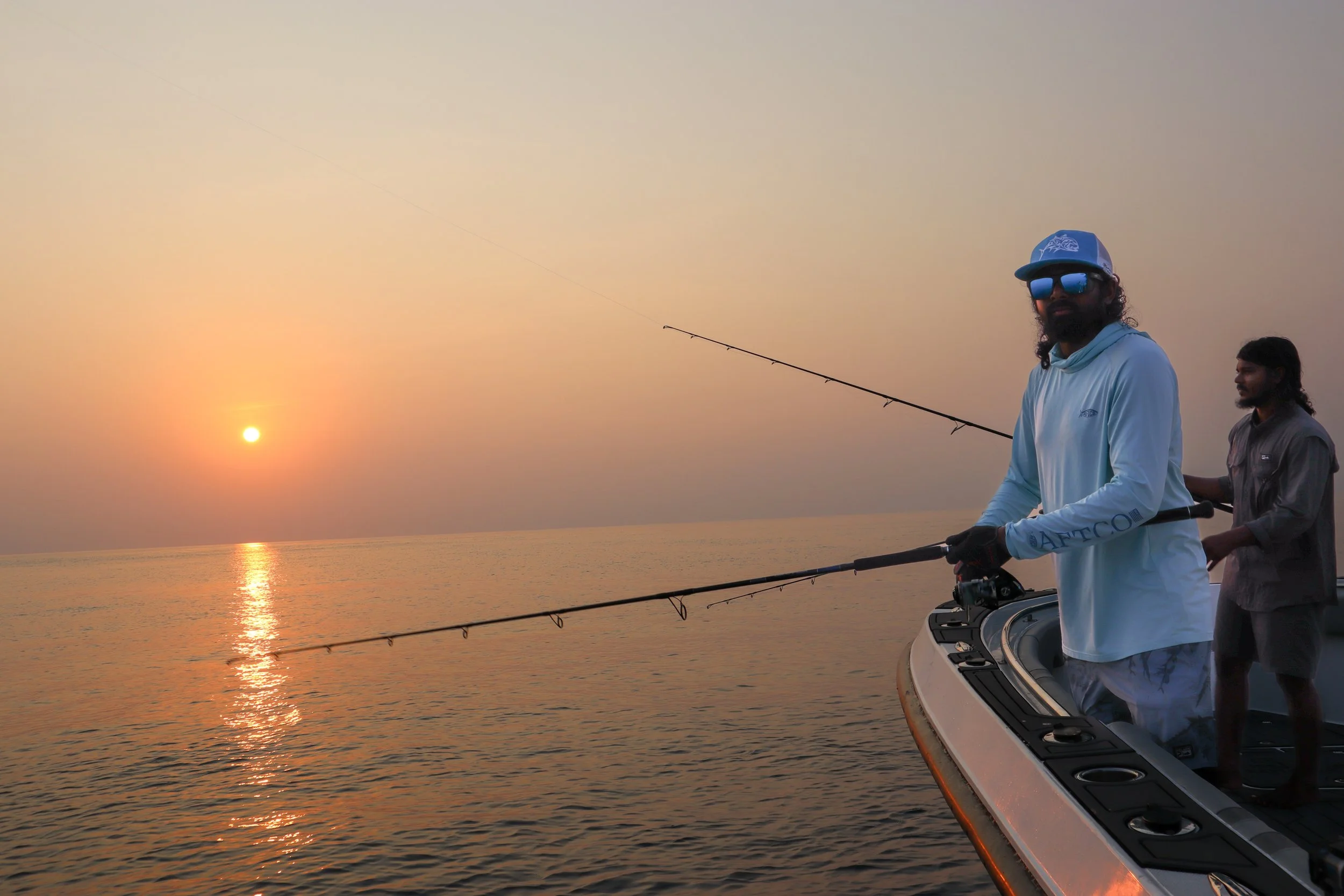 Two men fishing off a boat during sunset on the water, with one man wearing sunglasses and a cap.