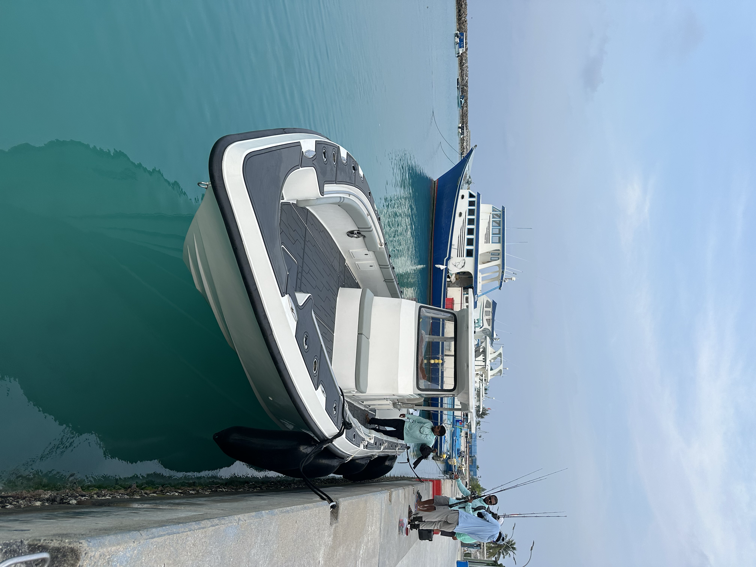 Motorboat docked at marina with two men fishing nearby, multiple boats in water, overcast sky.