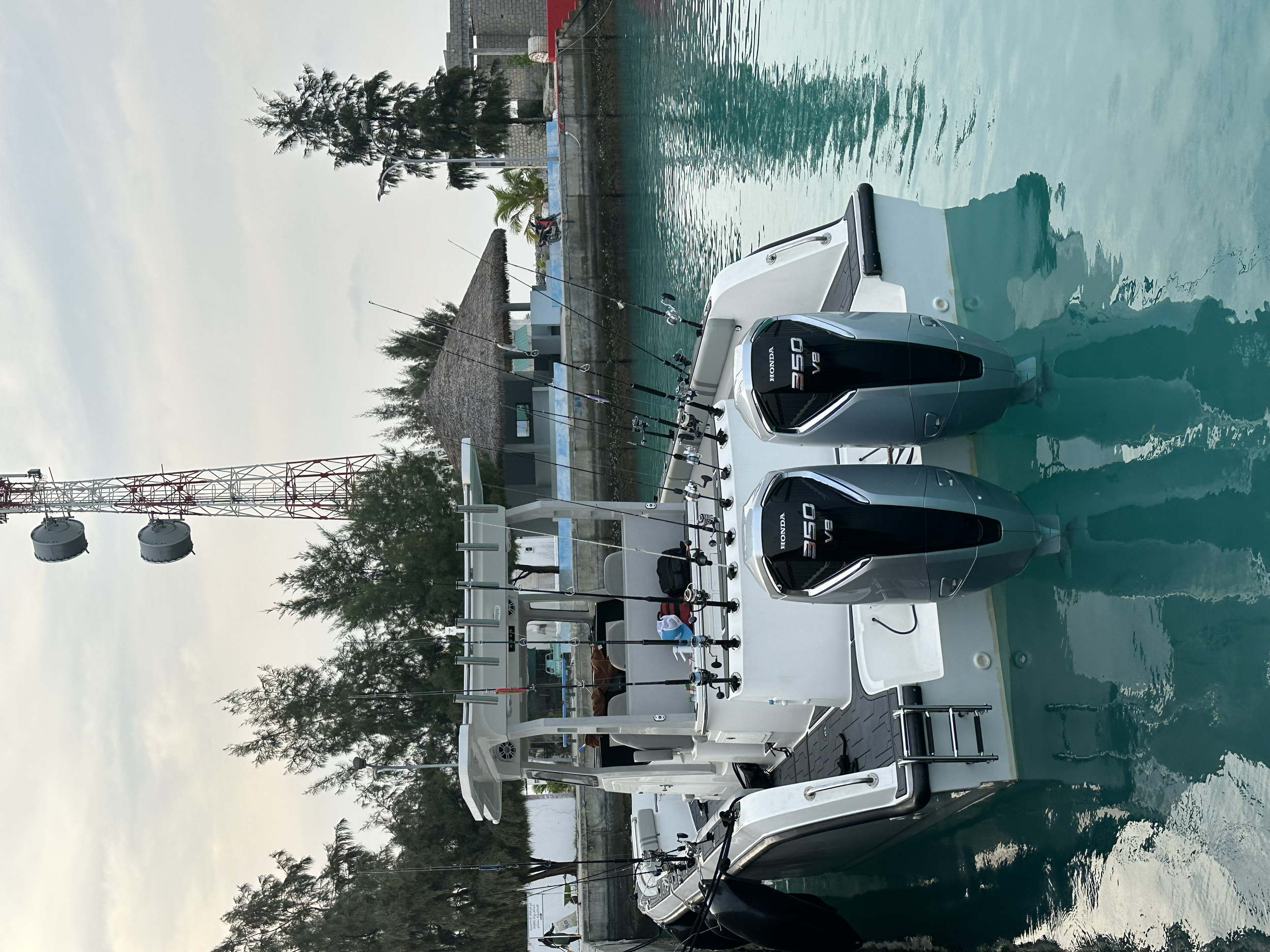Motorboat with two Honda 350 outboard engines, docked in a marina with trees and a building with a thatched roof in the background.