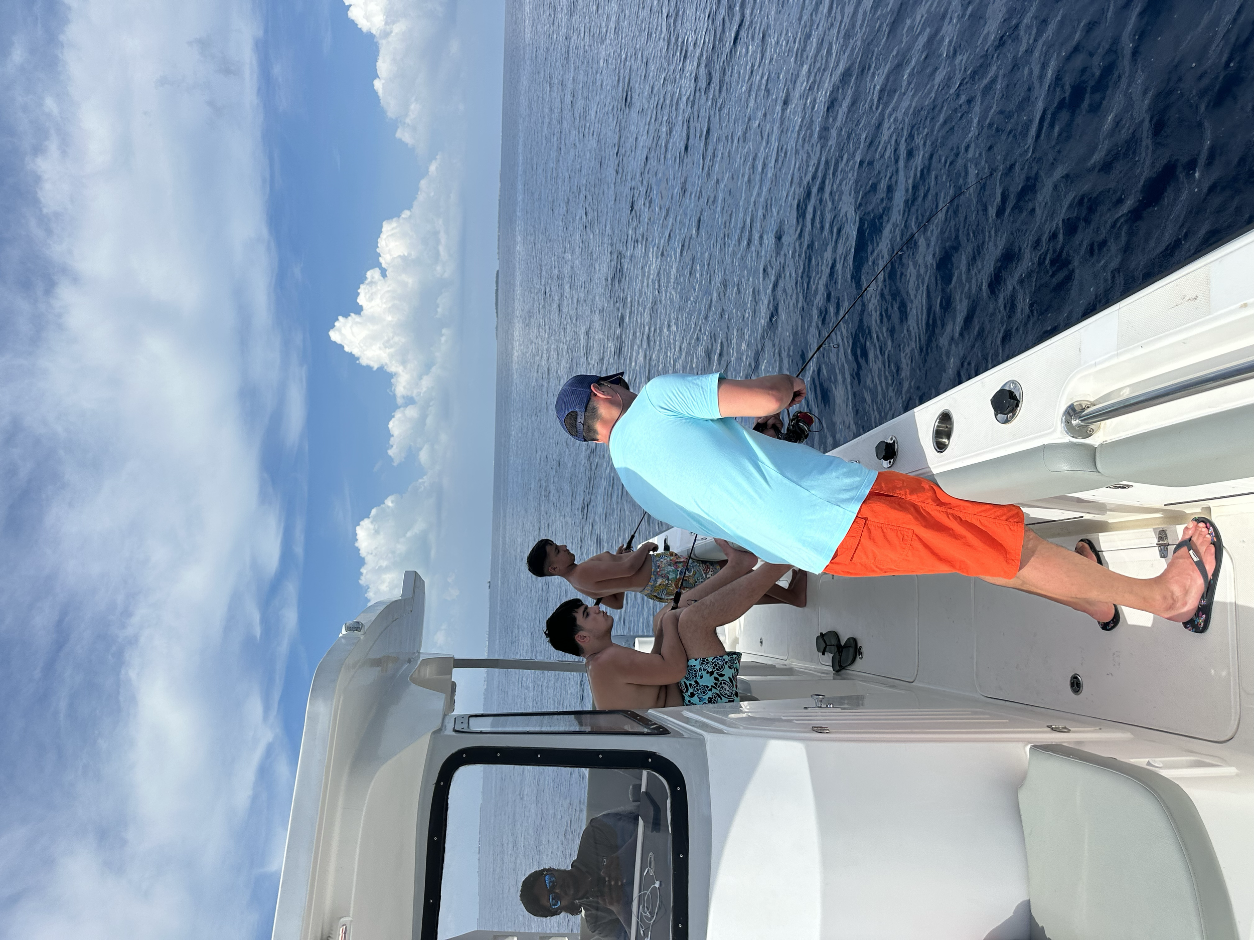 Four people on a boat, two sitting on the deck and two standing, one fishing into the water under a blue sky with clouds.