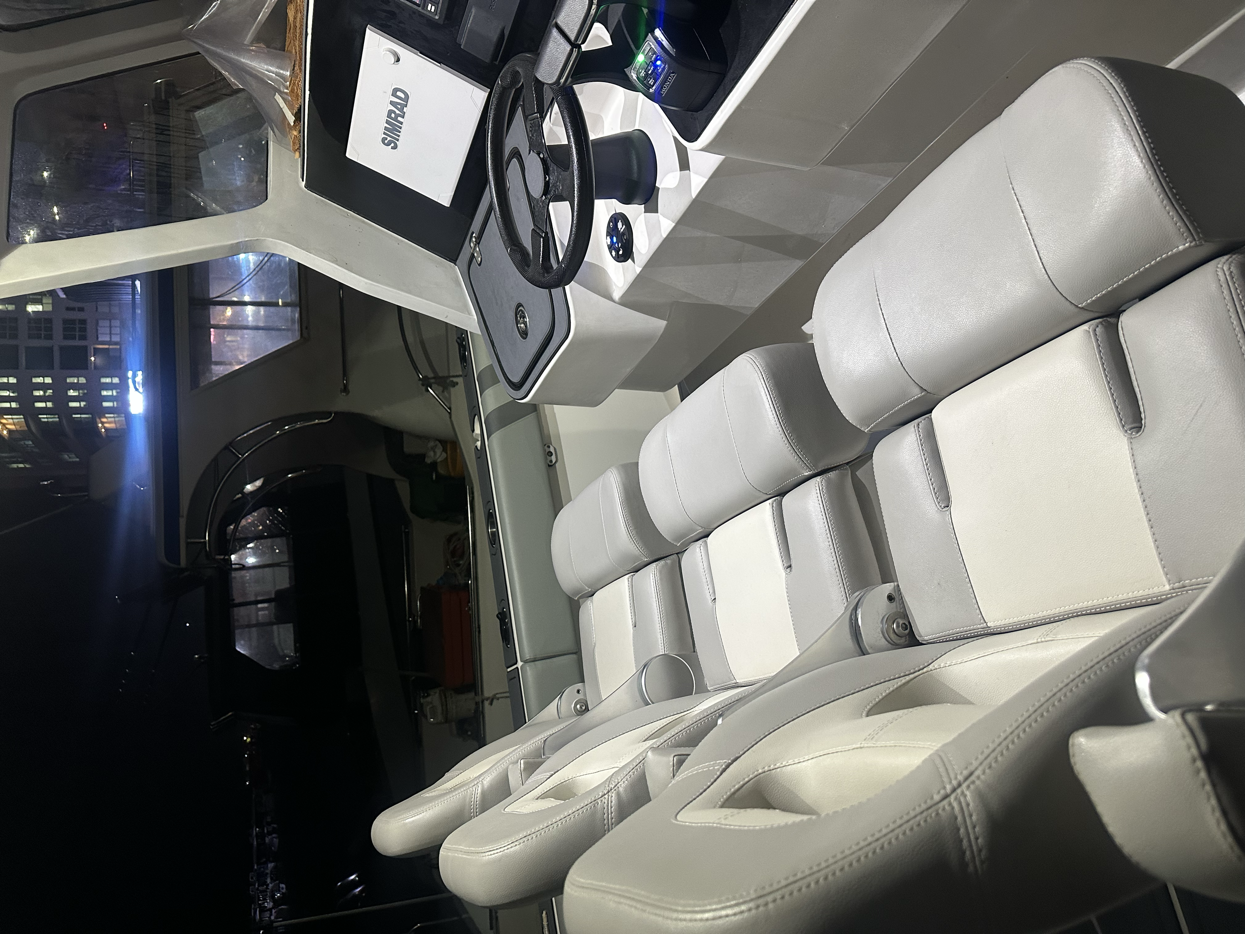 The interior of a boat cockpit showing a steering wheel, control panel, and white leather seats with a view of the city at night through the window.