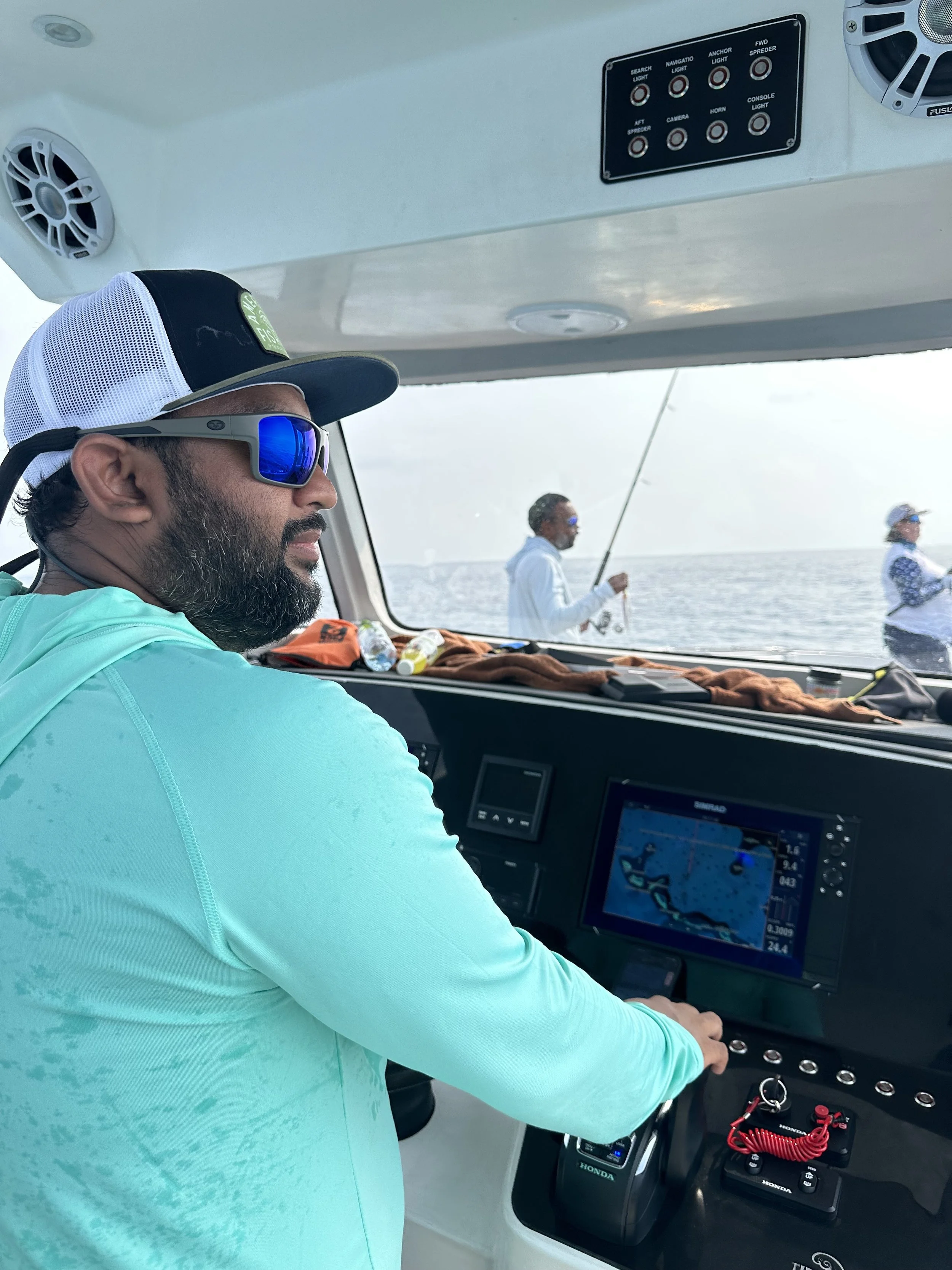 Man in boat cockpit with electronic navigation system, two men fishing in background, ocean view.