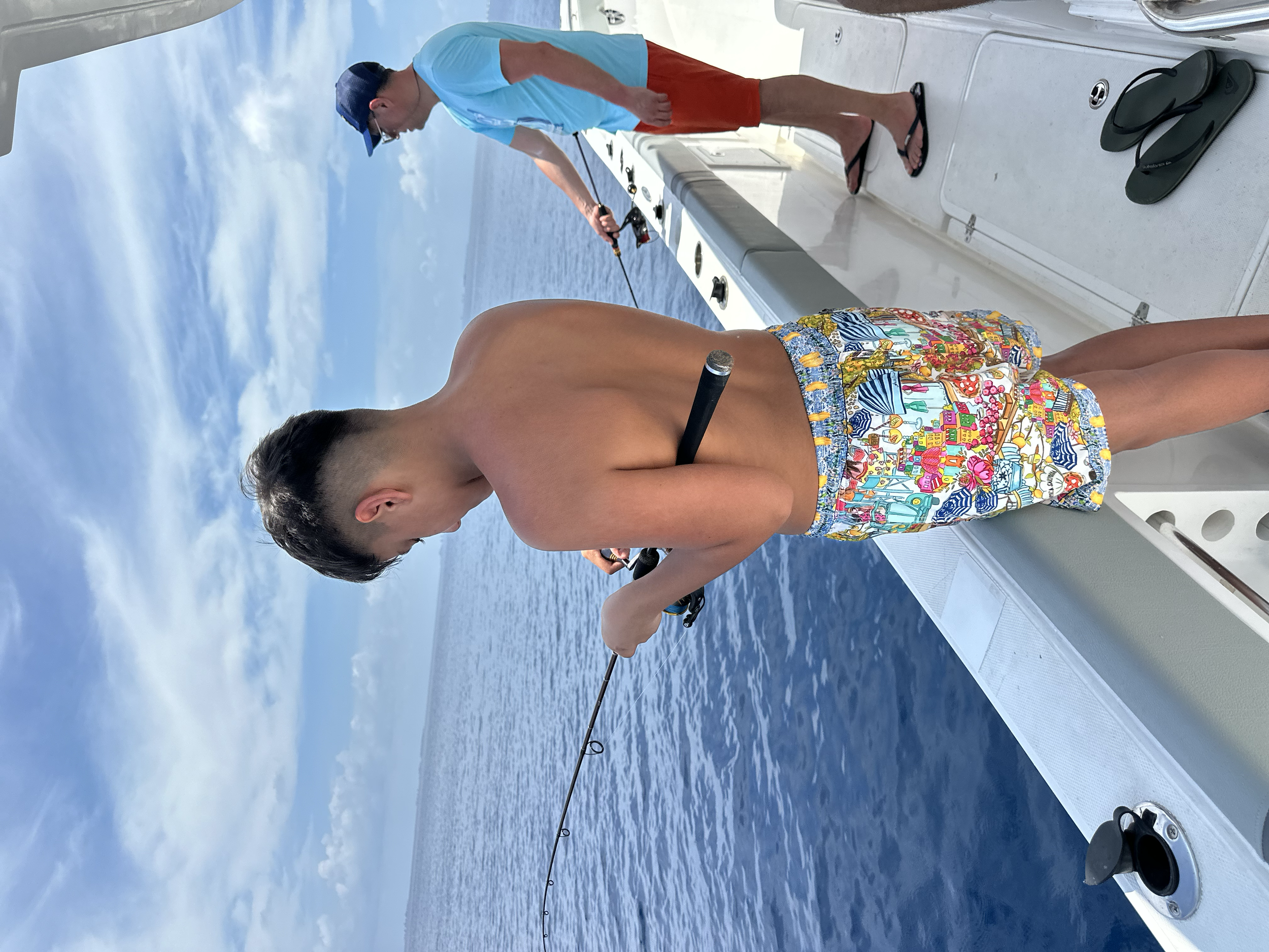 Two young men fishing off a boat on the ocean, with one in colorful swim trunks and the other in a blue shirt and shorts,Under a partly cloudy sky.