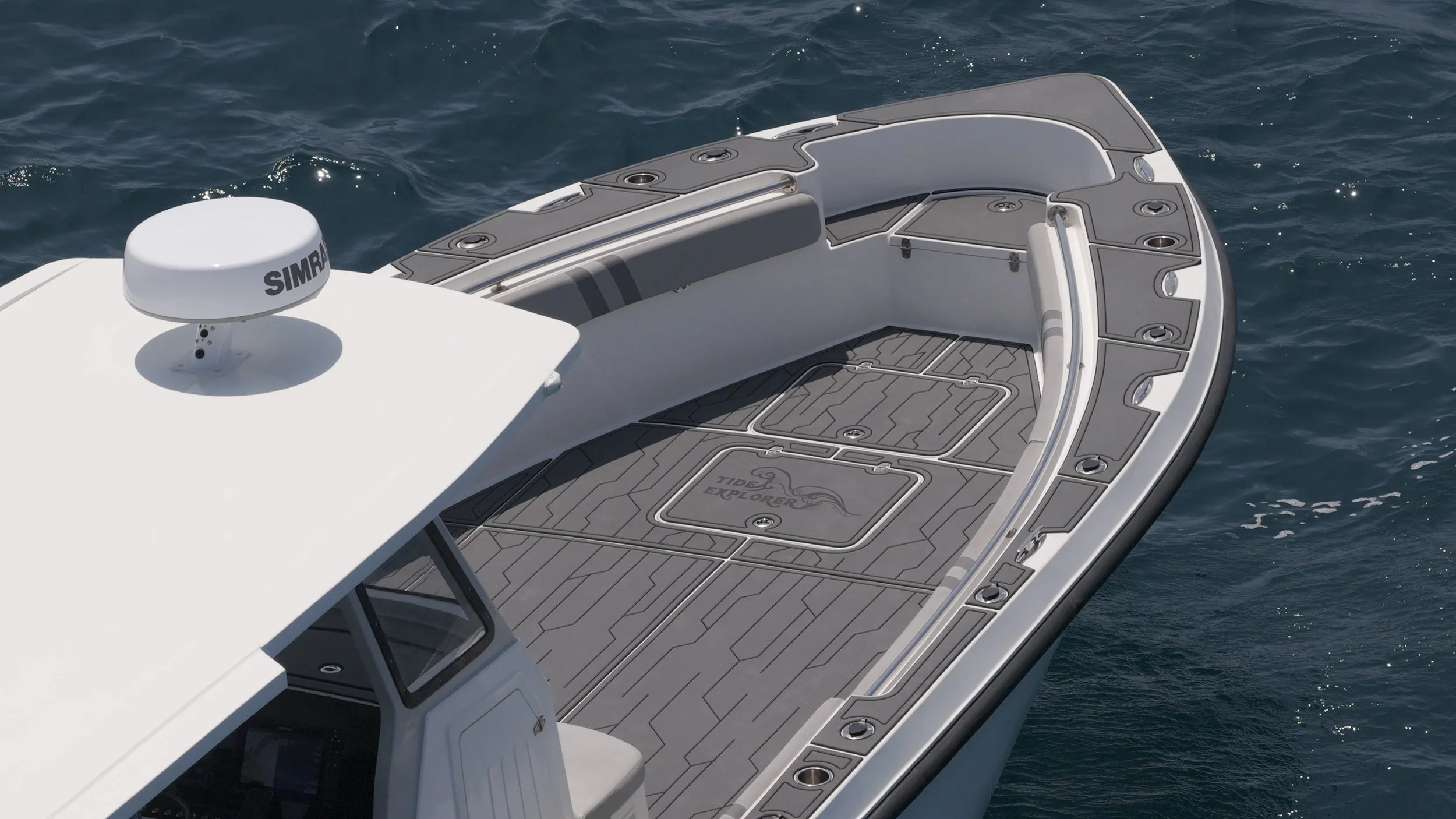 Close-up of the front section of a white yacht with a gray deck, ocean water in the background, featuring hatch covers, a small sink, and a radar with the brand name 'Simrad' on top.
