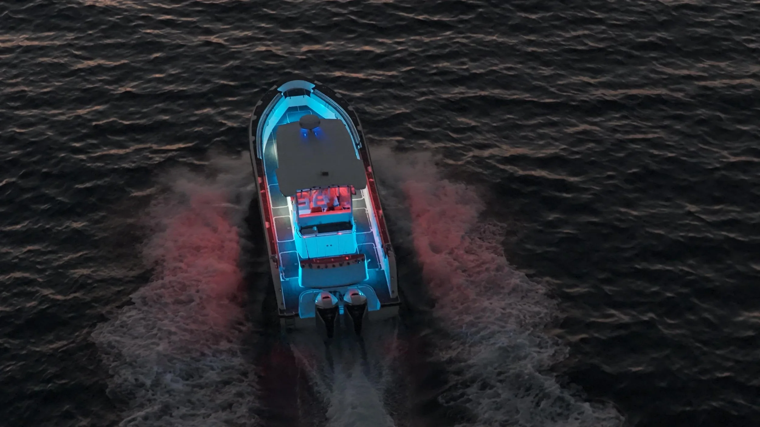 A boat with bright LED lights in blue and red colors seen from above, moving across dark water at dusk or night.