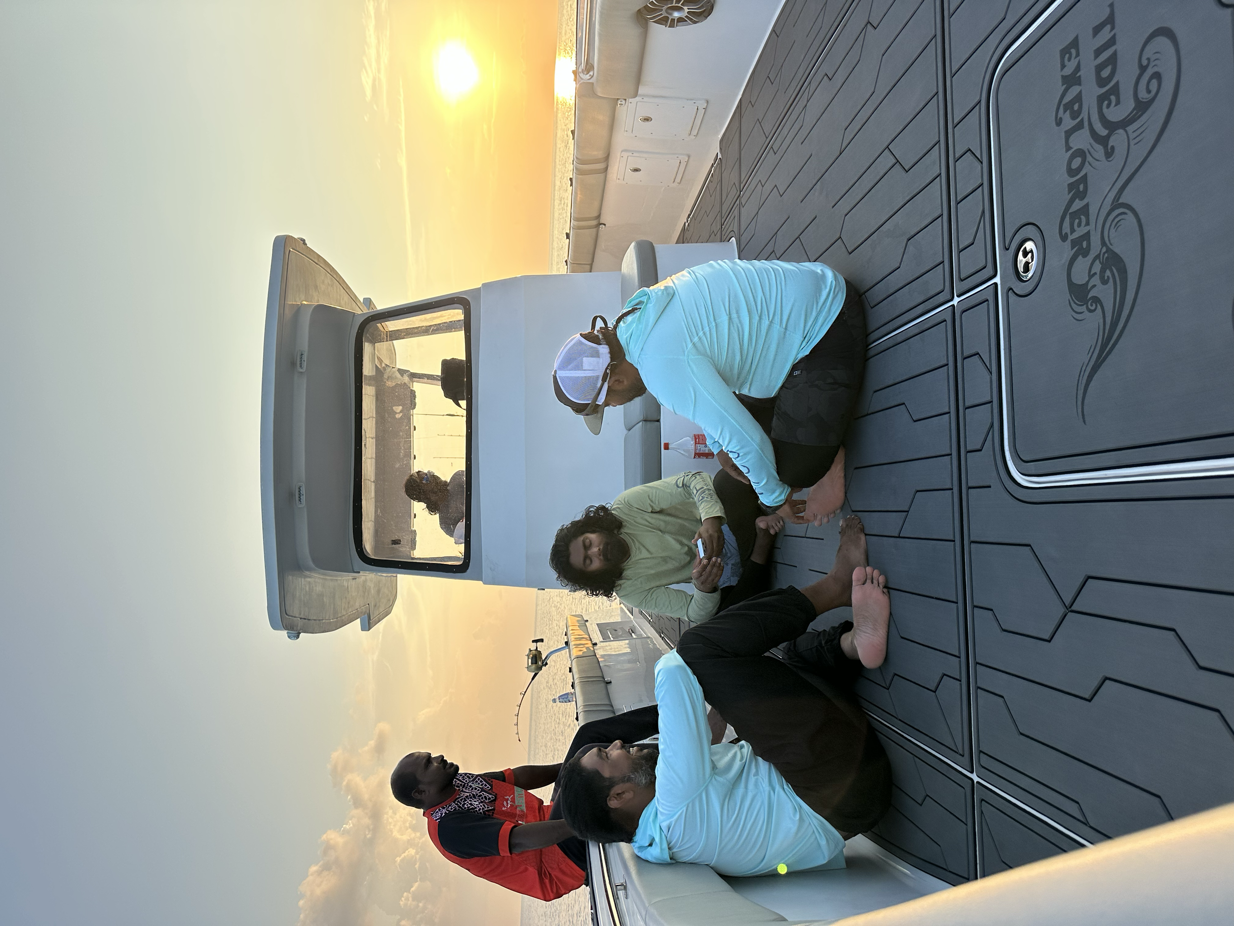 Group of people sitting and standing on a yacht deck during sunset, with a woman using a smartphone and a man tying his shoe.
