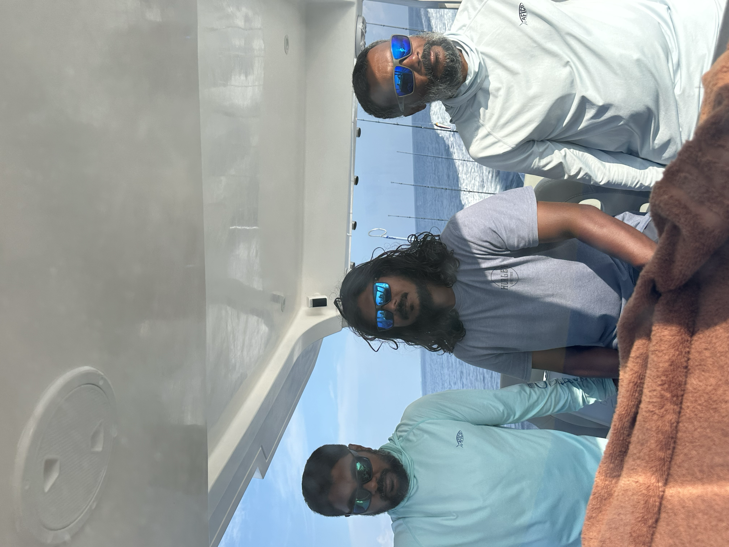 Four men on a boat with the ocean and a blue sky in the background, all wearing sunglasses. One man has long wavy hair and a beard, another has short hair and a beard, one has a beard and short hair, and the fourth man has short hair and is wearing a