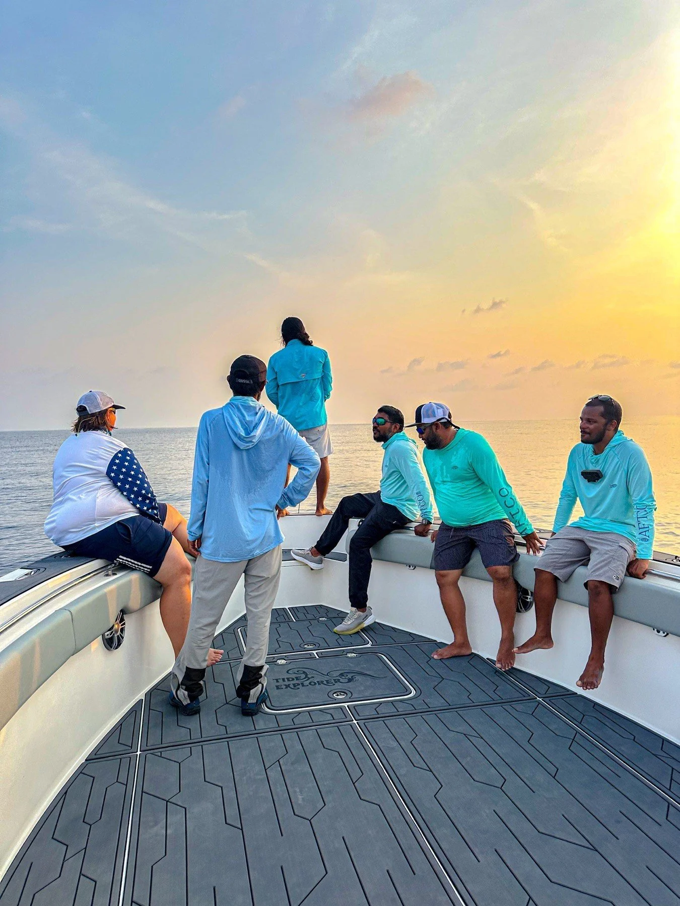 A group of five people on a boat during sunset, with some sitting and others standing by the railing, looking out at the water and the sky.