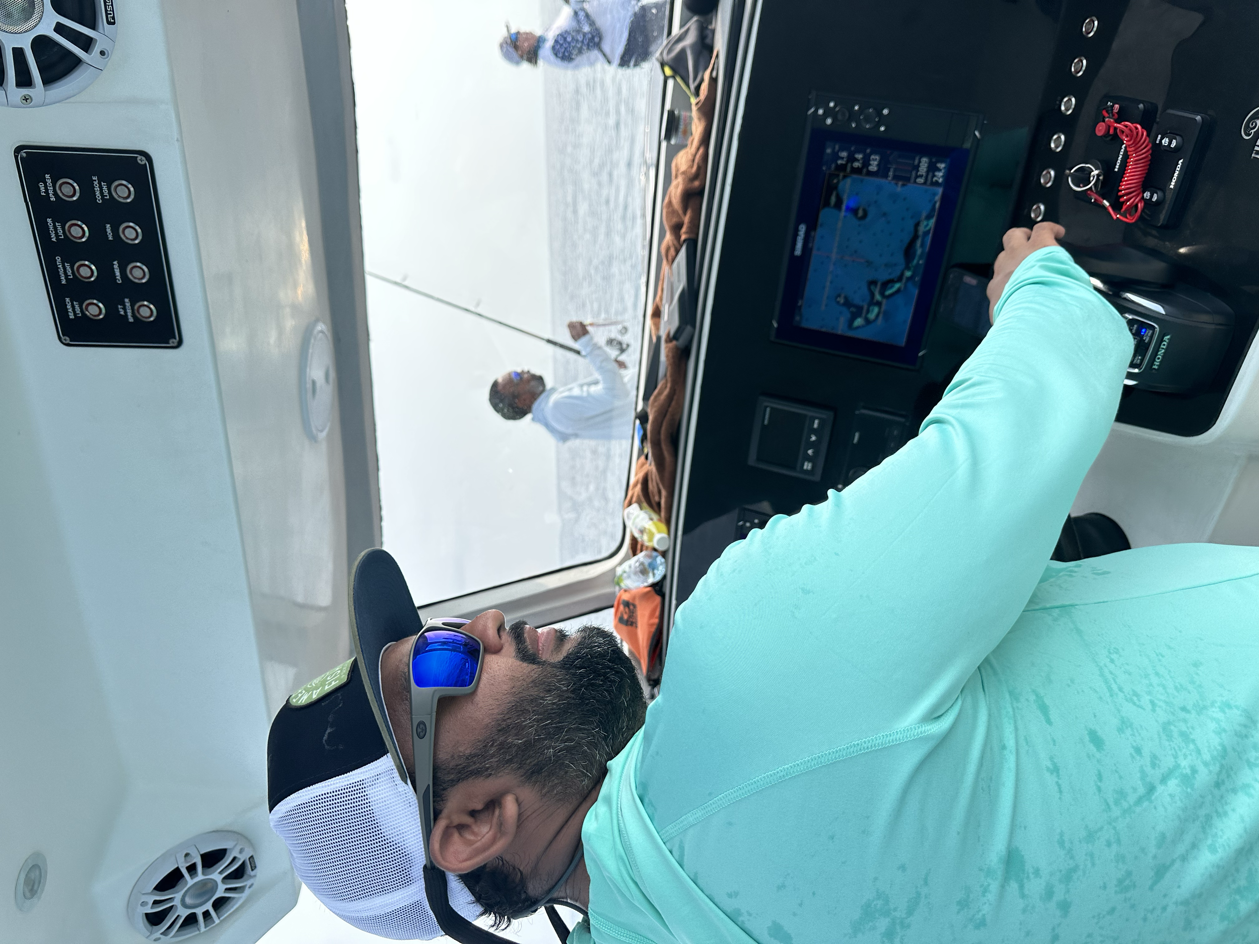 A man wearing sunglasses, a black and white cap, and a light blue shirt is operating a boat's navigation system. Two people are fishing off the boat in a foggy environment.