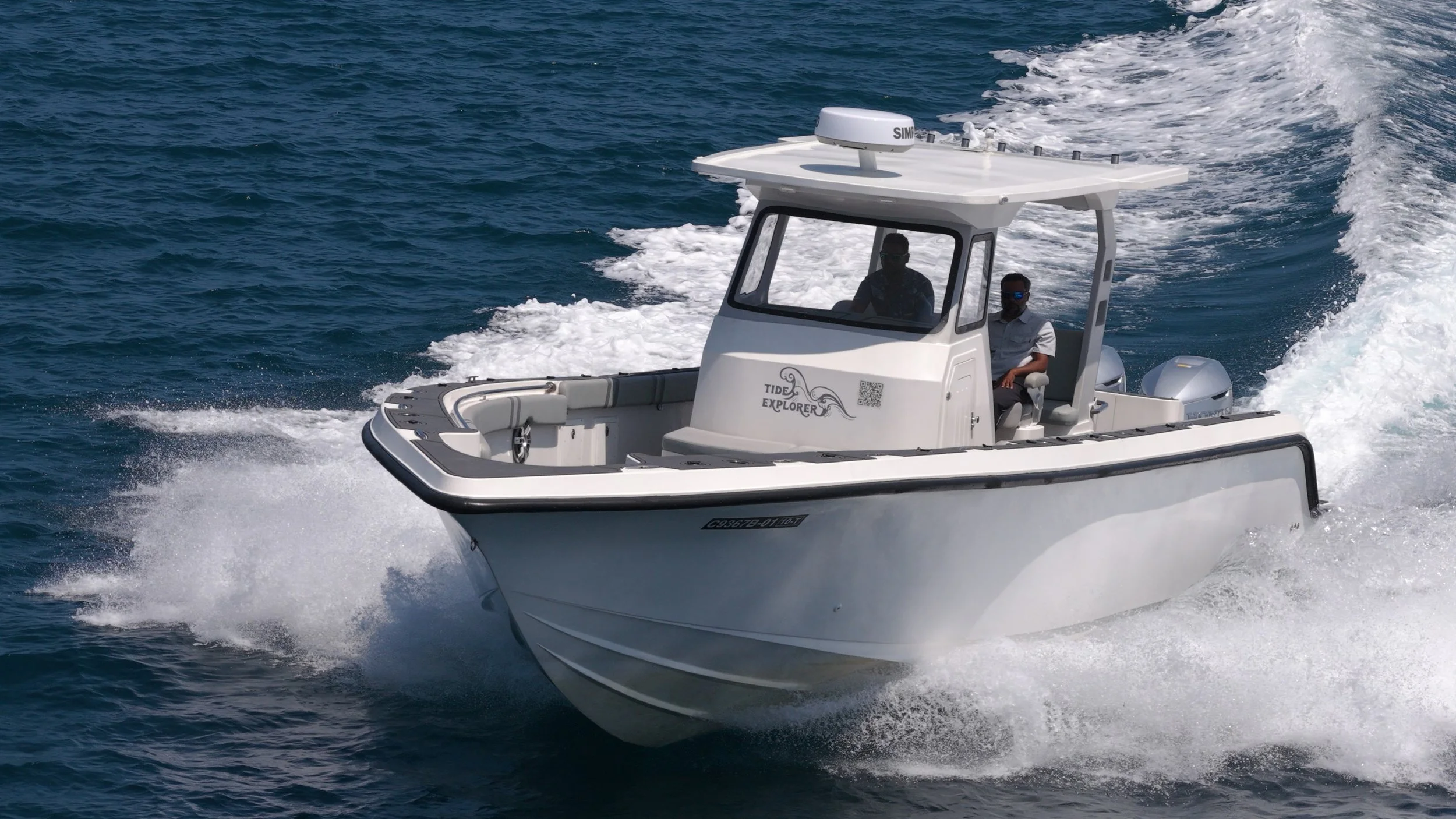 A white motorboat named Tide Explorer moving swiftly on blue water, with two people onboard wearing sunglasses.