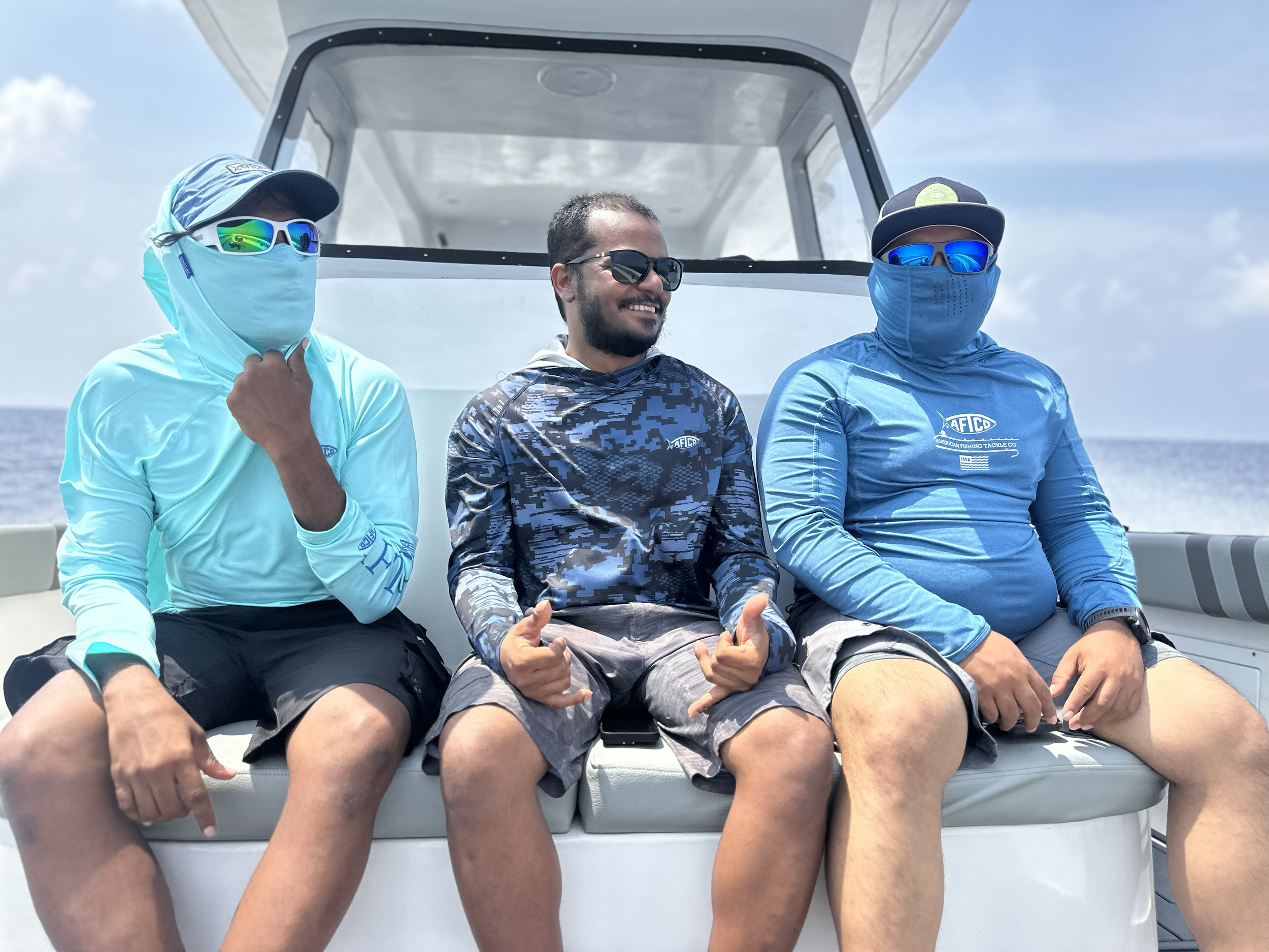 Three men sitting on a boat, all wearing sunglasses and sun protective clothing, with two of them giving thumbs up and smiling.