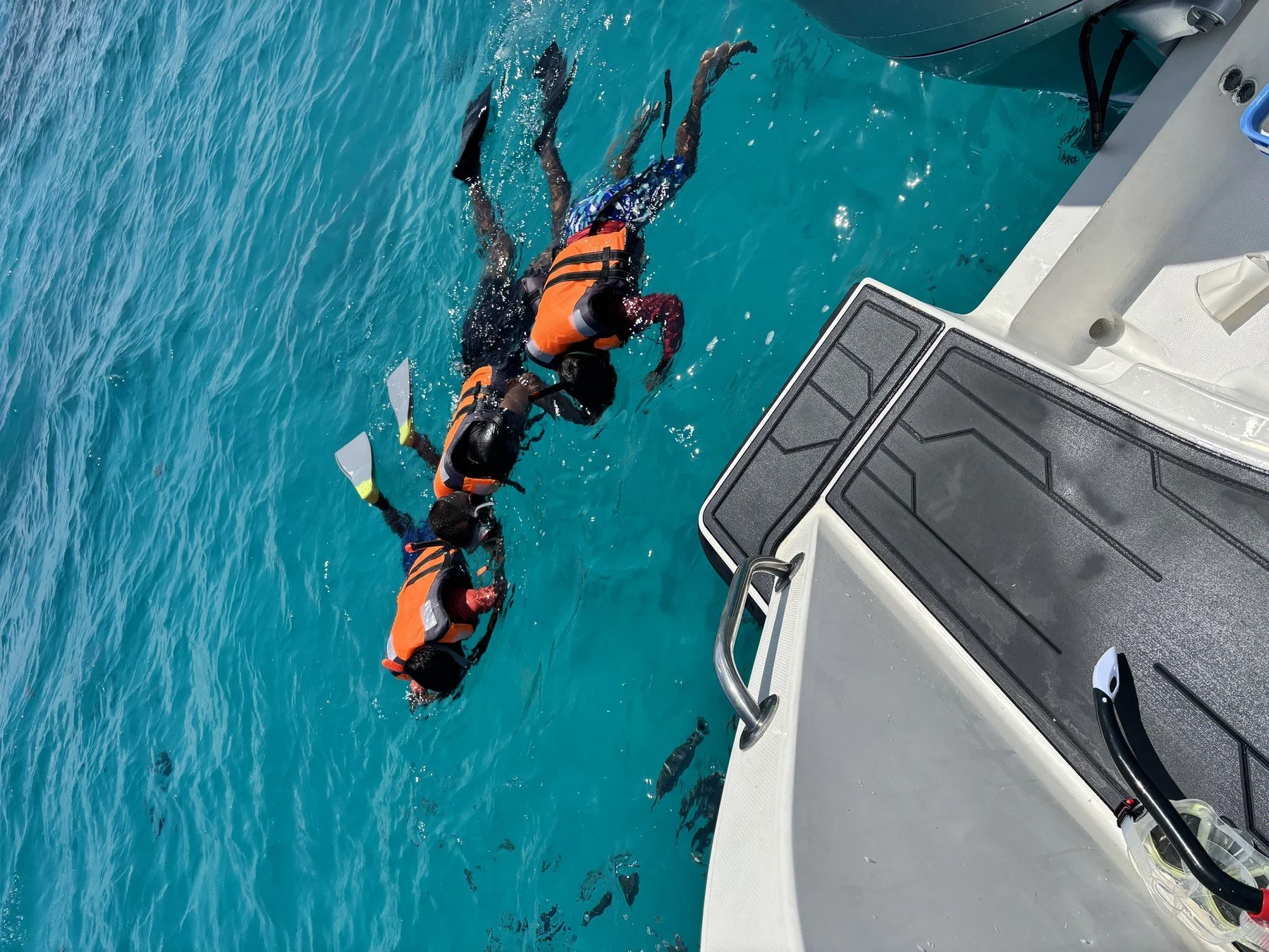Two people in orange life jackets swimming in clear blue water near the side of a boat.
