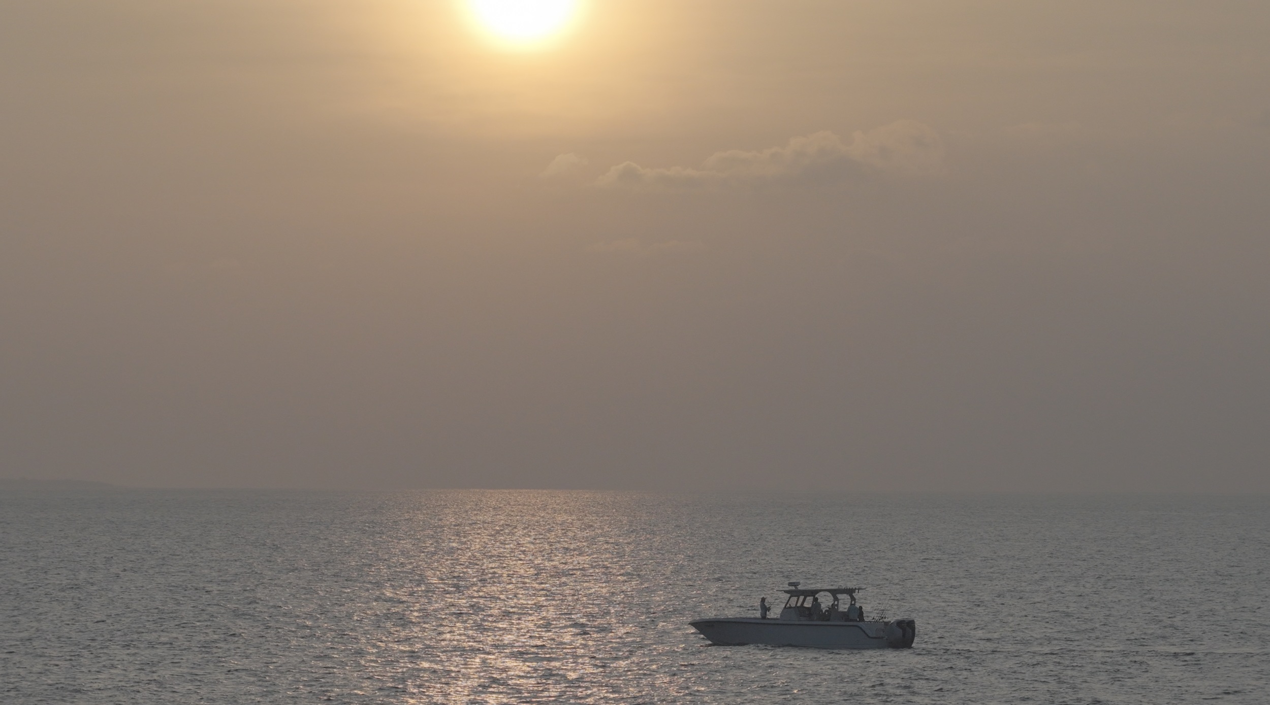 A boat on the ocean near the horizon with the sun partially obscured by clouds, creating a hazy glow over the water.