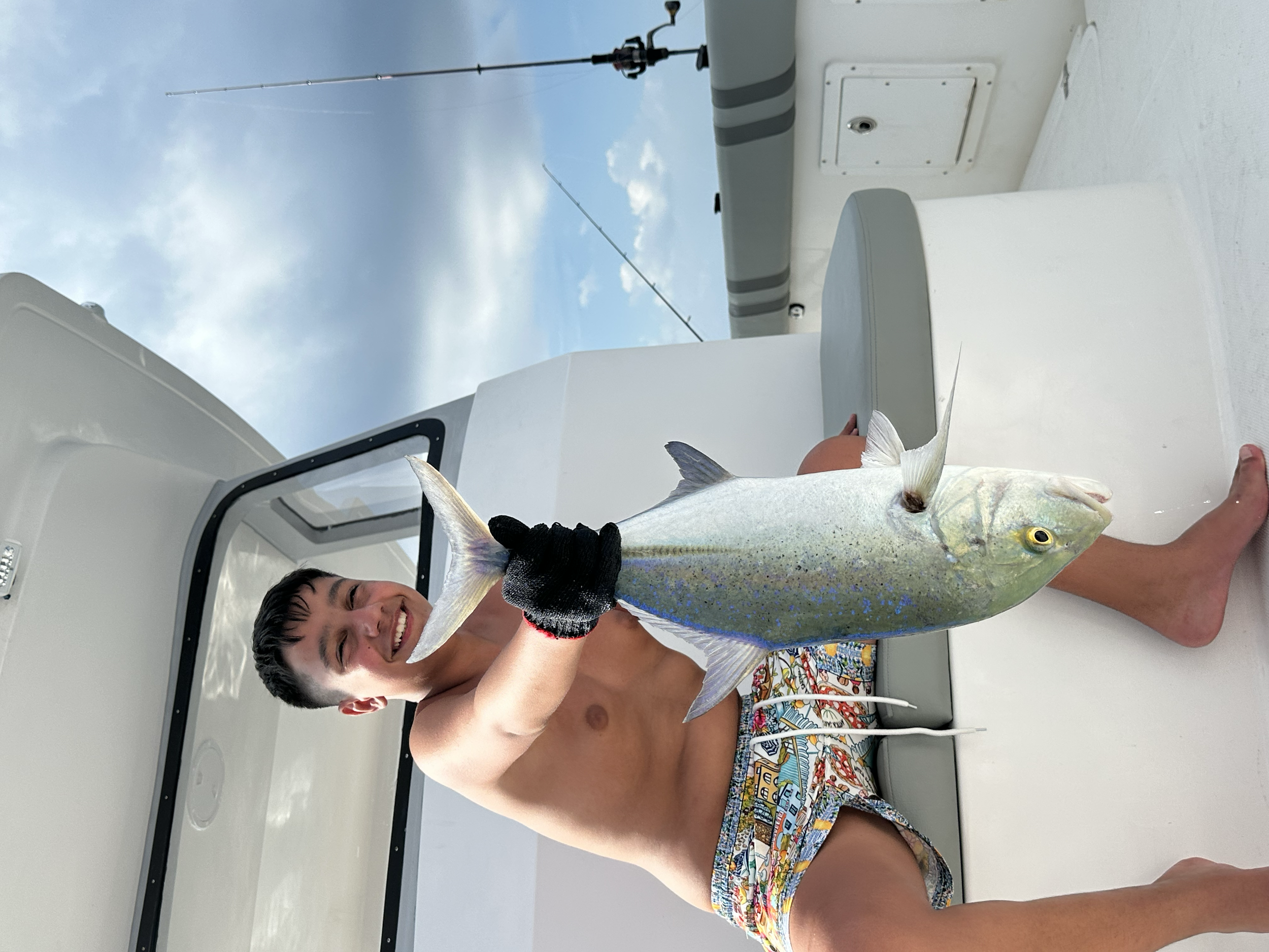 Smiling young man in colorful shorts holding a large caught fish on a boat with fishing rods and cloudy sky in the background.