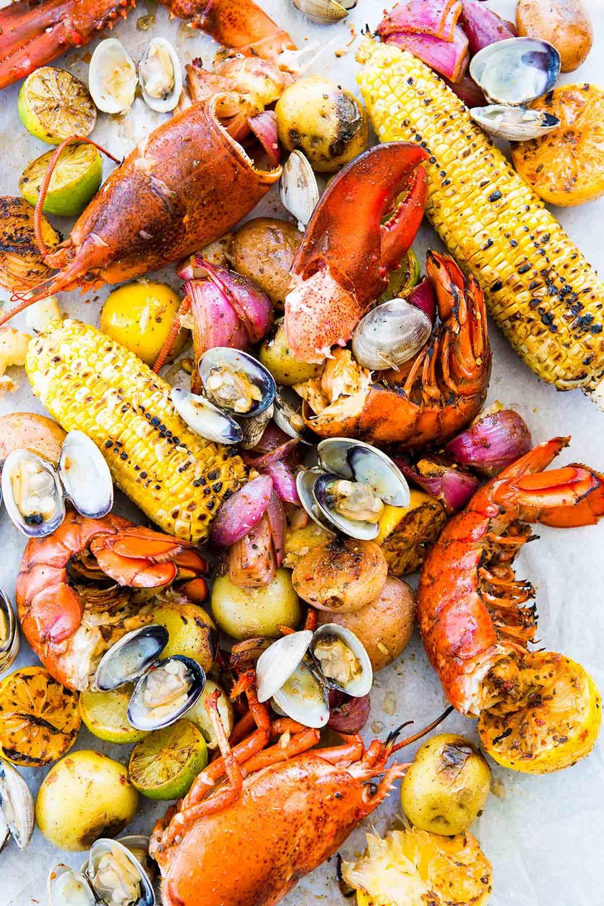 A variety of cooked seafood including lobster, shrimp, clams, corn on the cob, potatoes, and onions, displayed on a white surface.