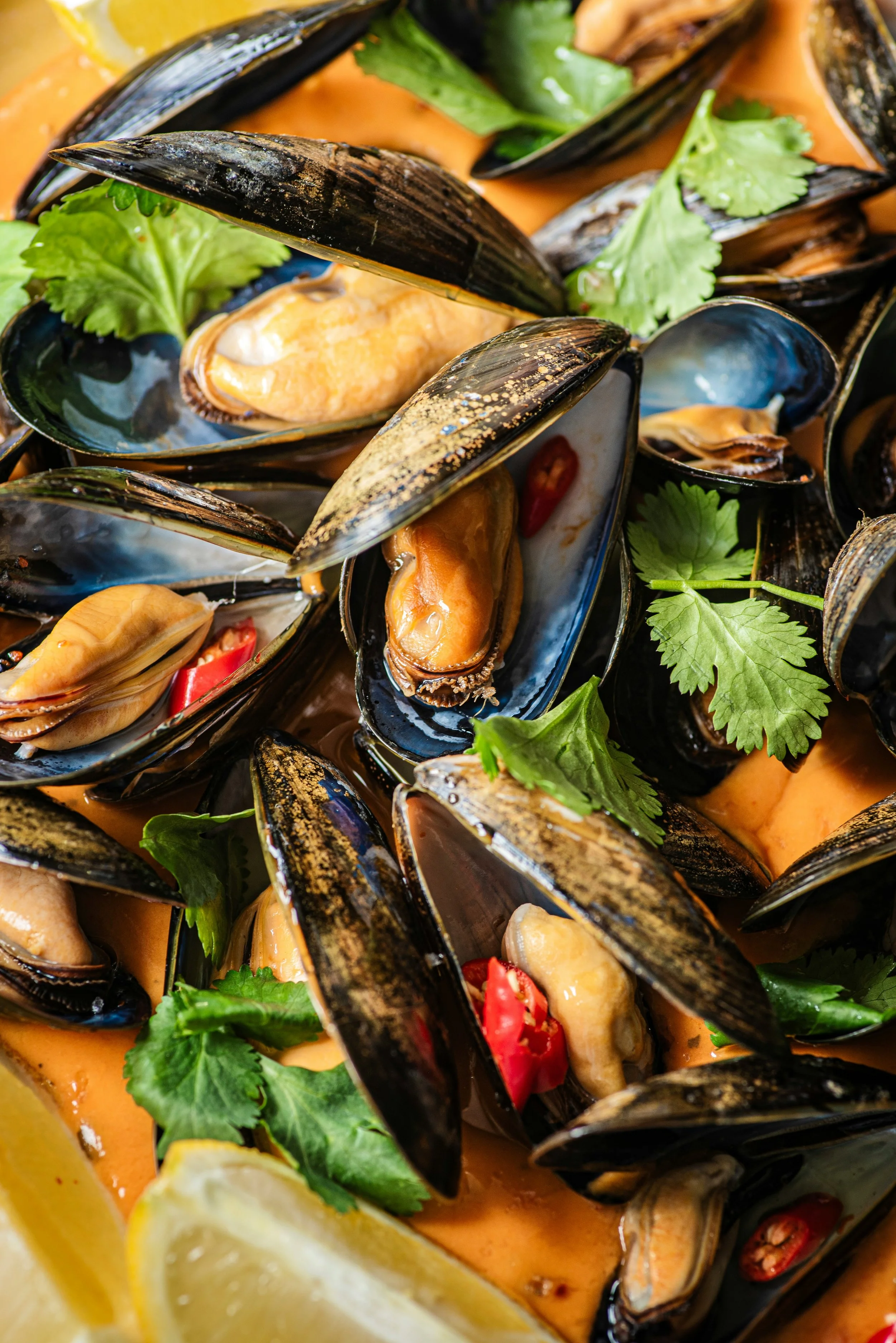 Close-up of cooked mussels in orange sauce with cilantro, red chili slices, and lemon wedges.