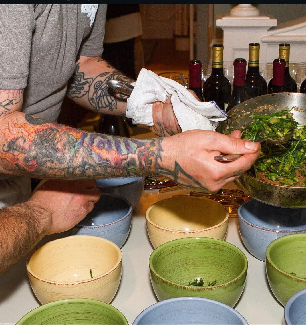 Person with tattooed arms serving cooked greens into bowls on a table at a gathering or event.