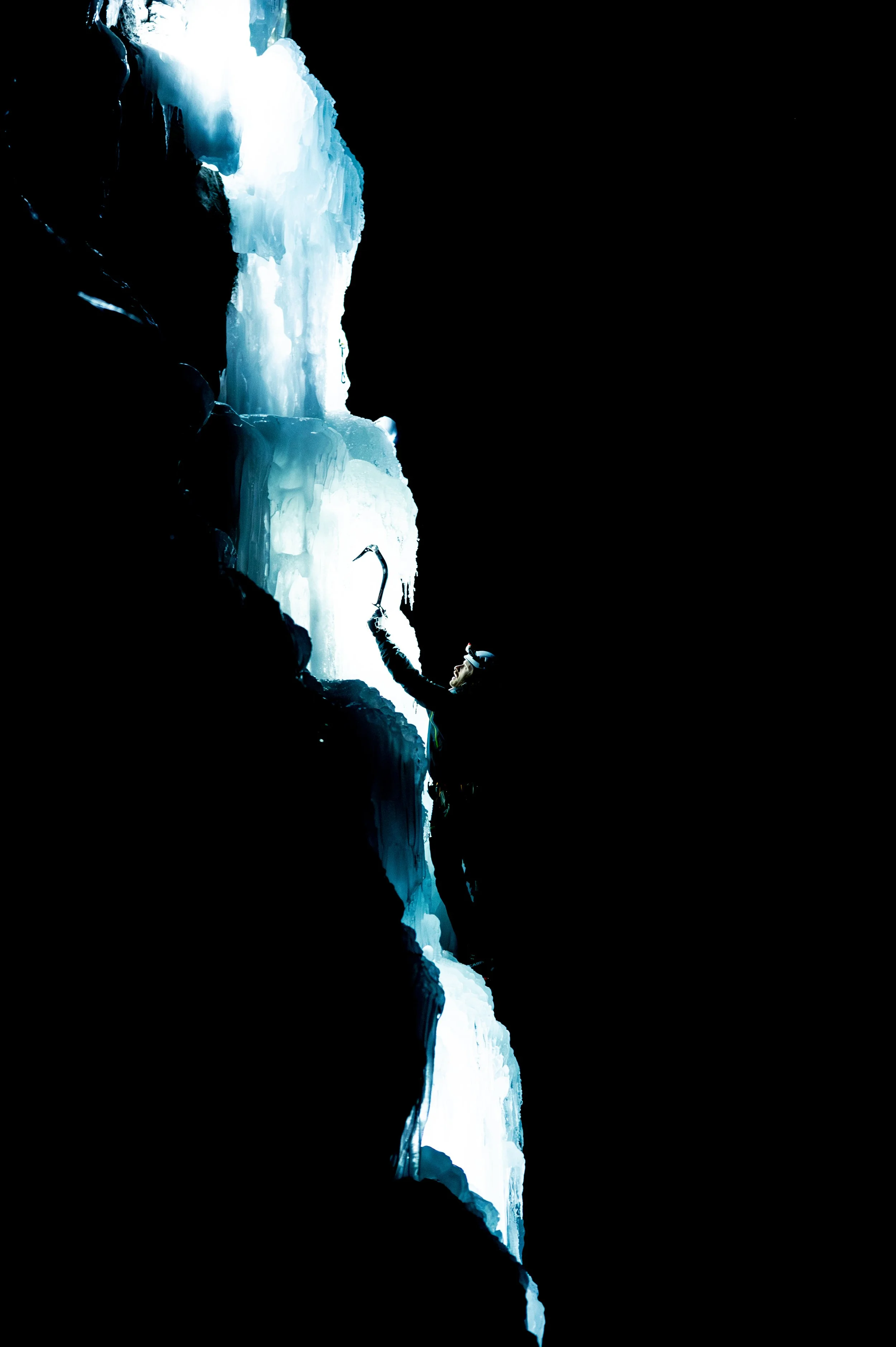 Un alpiniste escaladant une paroi de glace de nuit dans une grotte ou un canyon glacé, avec des formations de glace suspendues et une silhouette contrastée dans l'obscurité.