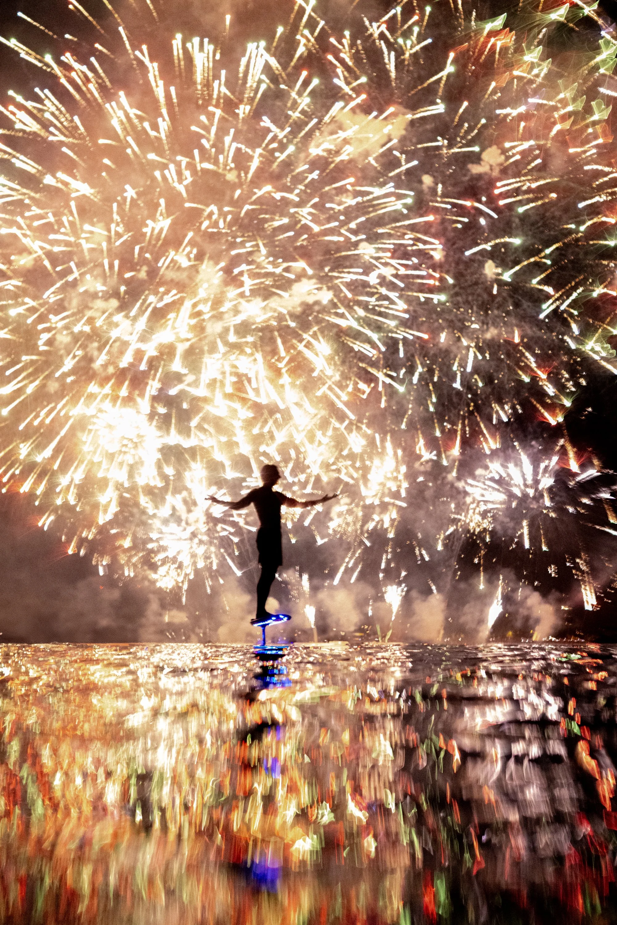 Une personne debout sur un hoverboard illuminé, avec des feux d'artifice éclatants dans le ciel nocturne, reflétés sur une surface humide.