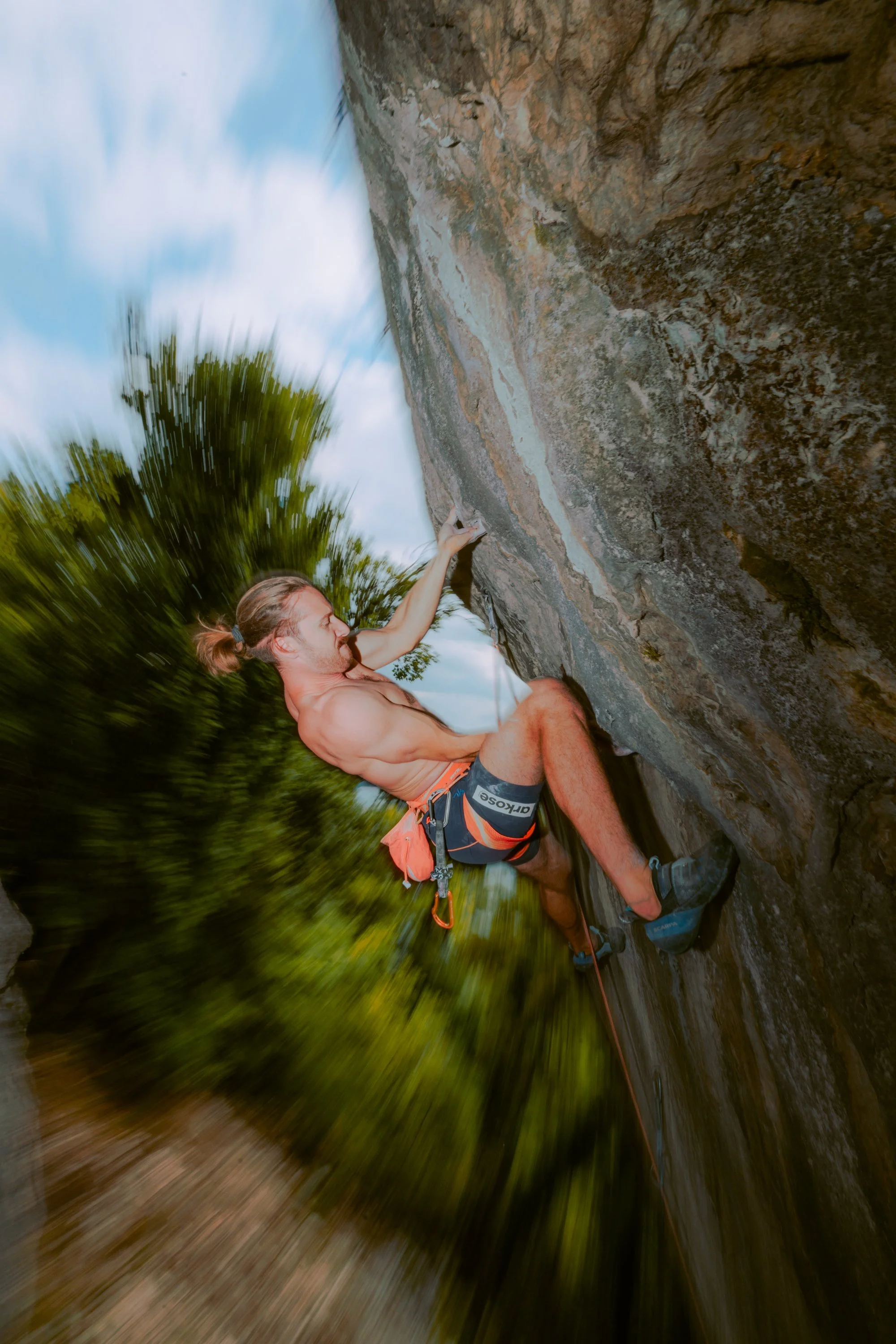 Un homme pratiquant l'escalade sur une falaise rocheuse en extérieur, sans chemise, portant un short et des équipements d'escalade.