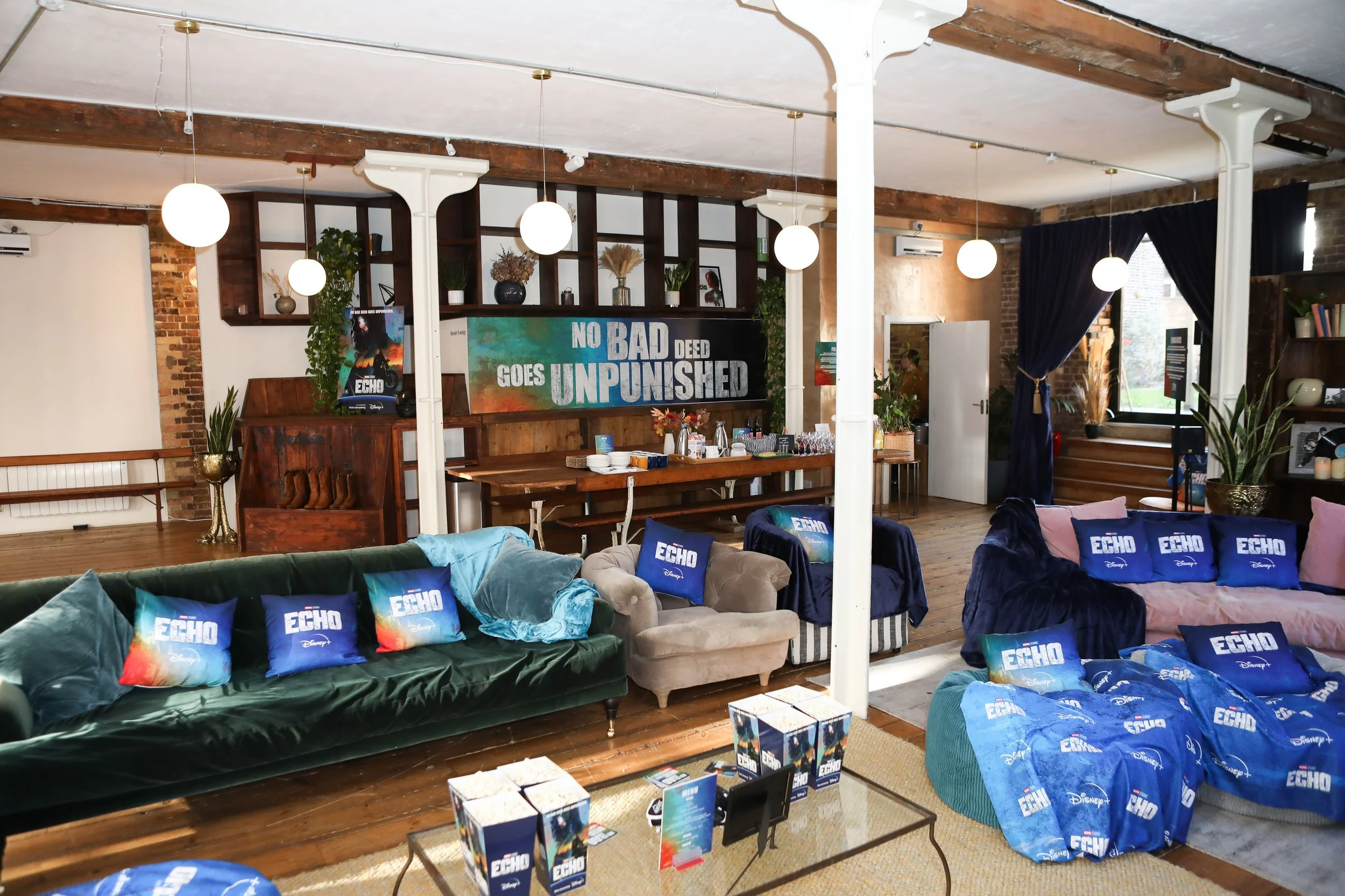 Living room decorated with promotional pillows and blankets for Disney's Echo, featuring sofas, chairs, a coffee table, and a large banner with the message 'No bad deed goes unpunished' filmed with plants and lighting.