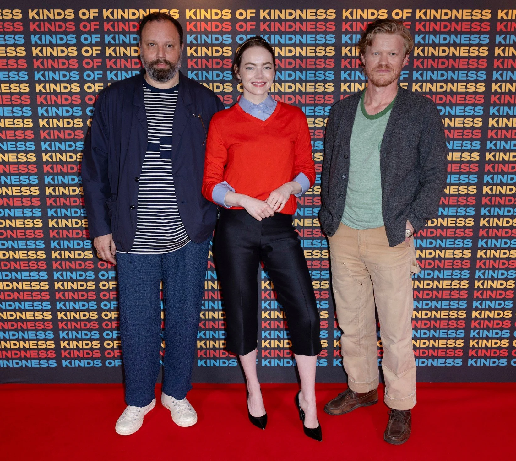 Three individuals standing on a red carpet in front of a backdrop with repeated text that says "KIDS OF KINDNESS" in yellow, red, and blue. The person on the left is a man with a beard, wearing a navy jacket over a black and white striped shirt, blue