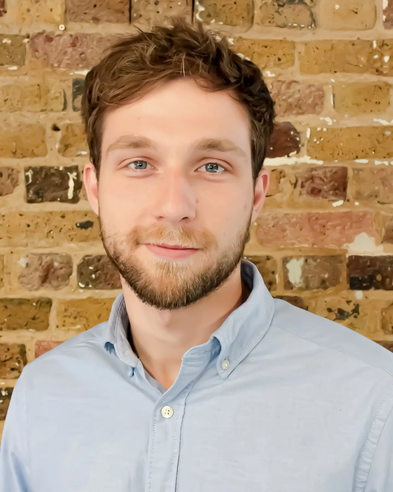 A young man with brown hair and a beard wearing a light-colored button-up shirt, standing in front of a brick wall.