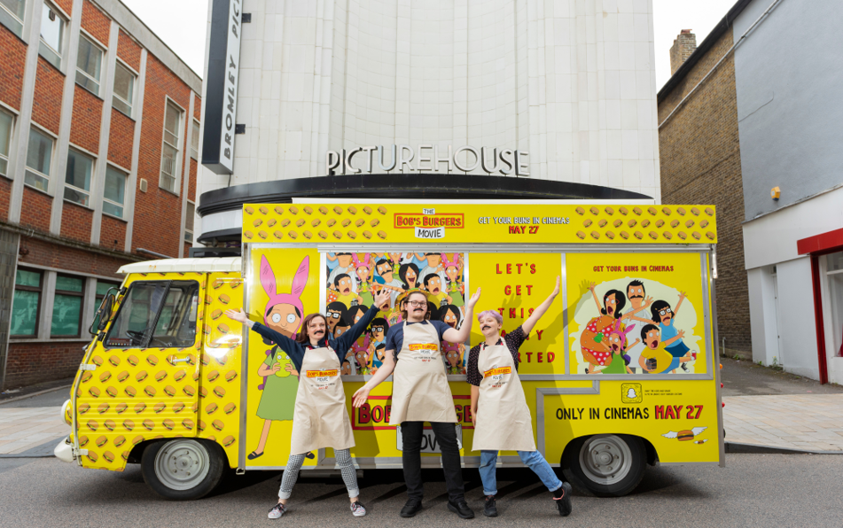 A brightly colored food truck with 'Bob's Burgers Movie' branding parked in front of the Picturehouse cinema, with three smiling people wearing aprons posing in front of it on a city street.