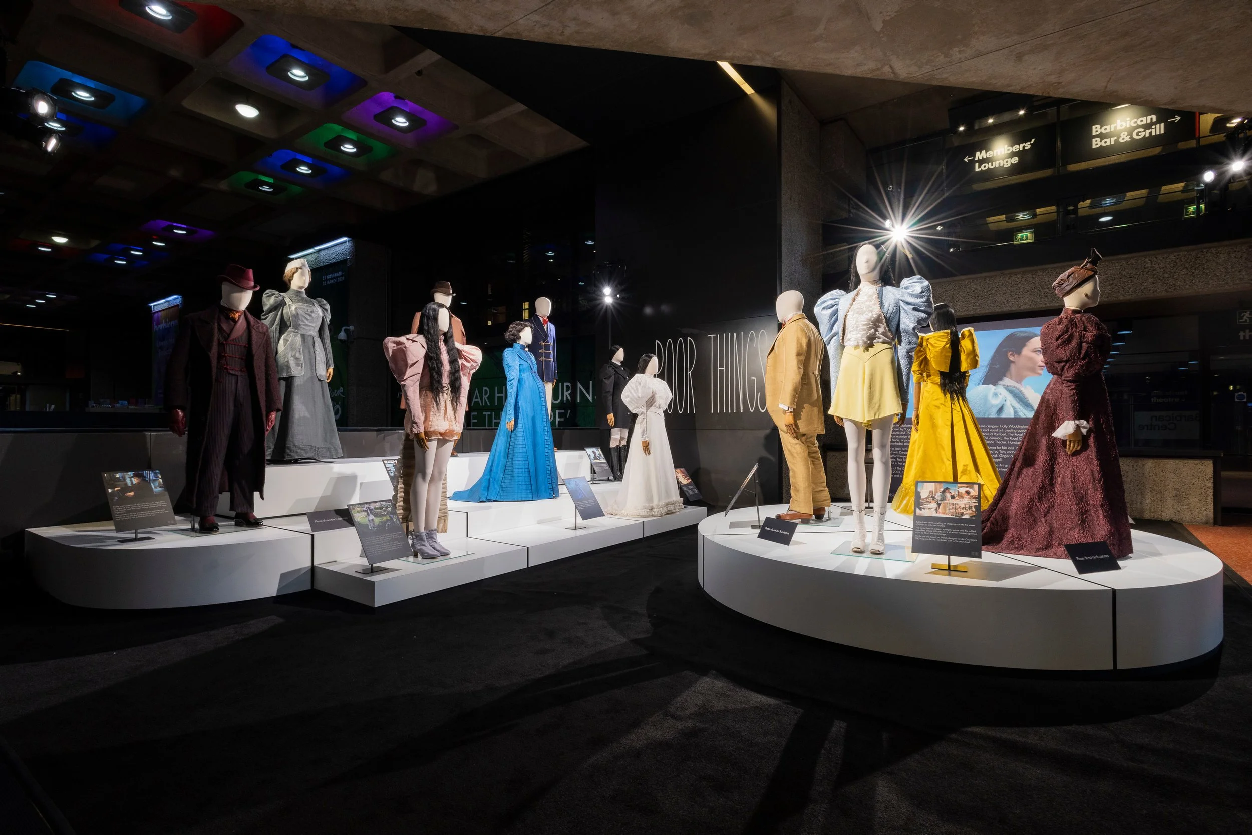 Display of mannequins dressed in vintage and historical costumes at a museum exhibit, with spotlights highlighting each outfit and informational plaques in front of some mannequins.
