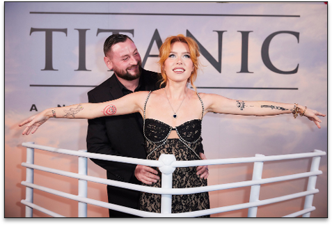 A woman with short orange hair and tattoos on her arms on a mock ship's railing, with a man behind her, in front of a Titanic-themed backdrop.