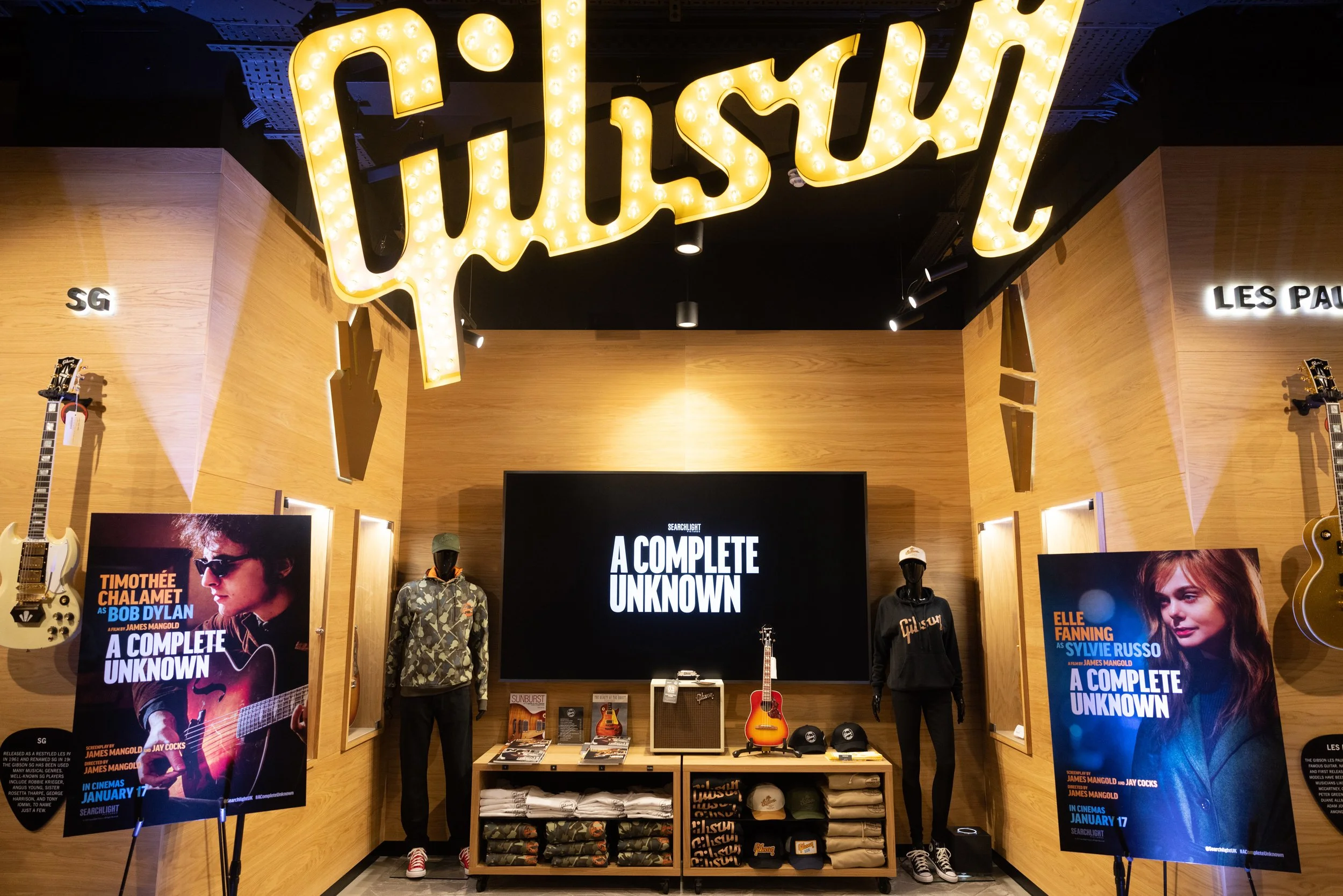 Display at a Gibson store with guitars on the wall, promotional posters for the movie "A Complete Unknown," mannequins wearing clothing, and a large illuminated Gibson sign overhead.