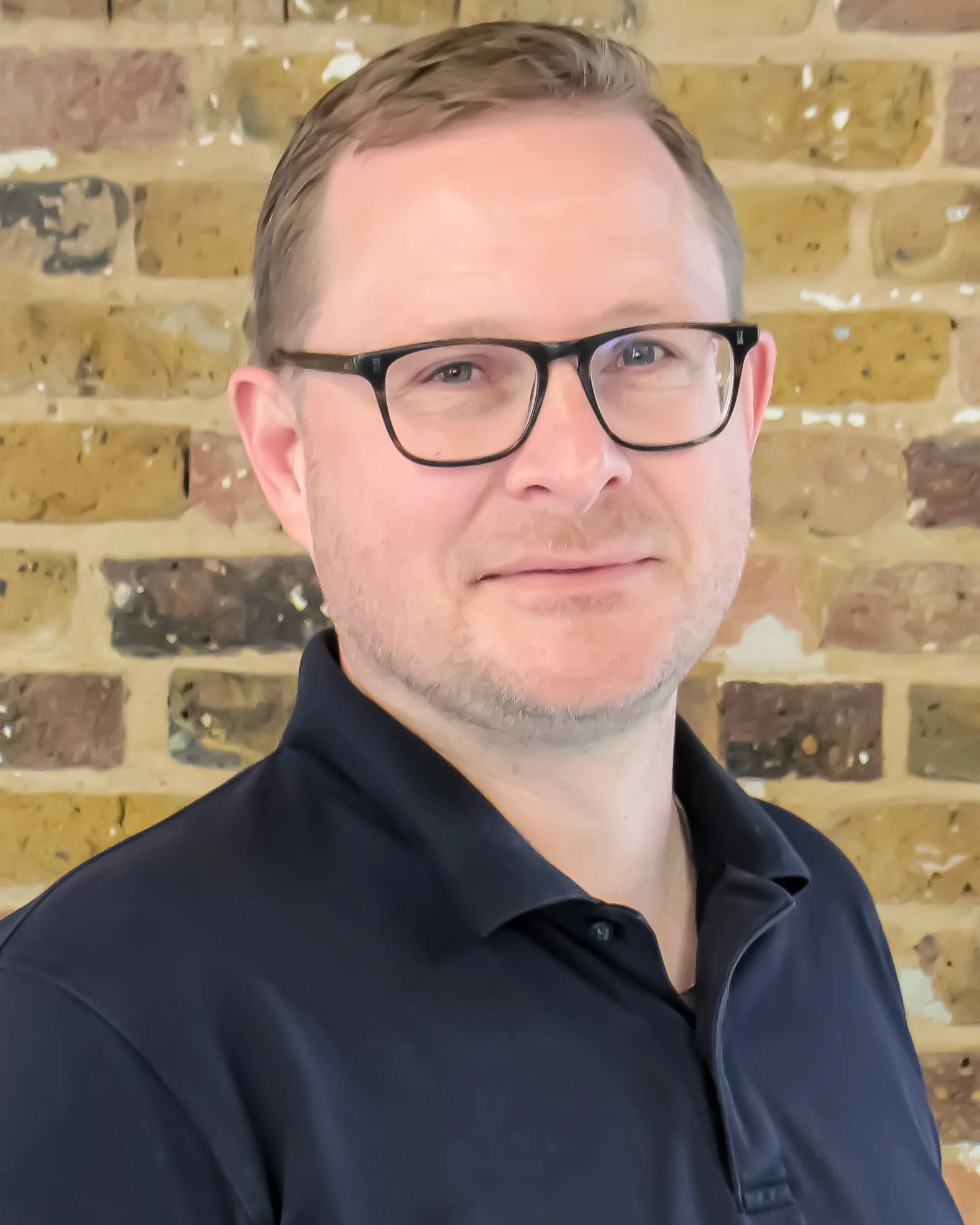 A man with short light brown hair, wearing black glasses and a black collared shirt, standing in front of a brick wall.