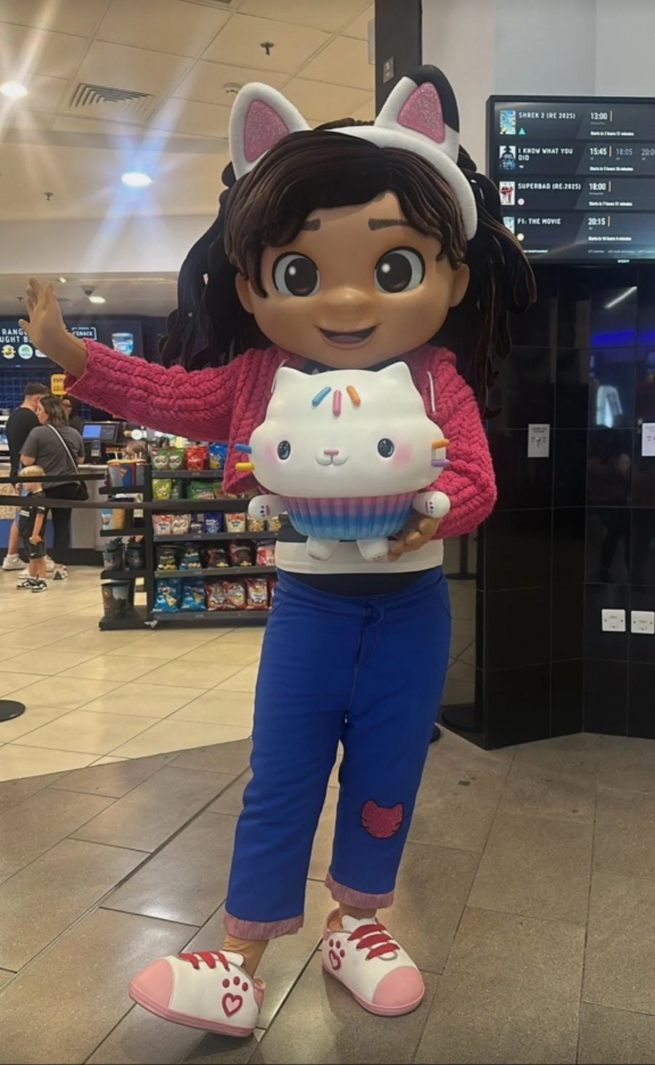 A person in a large cartoon-style character costume holding a cute cat-shaped plush toy, wearing a pink sweater, blue pants with a red cat patch, and shoes with heart and paw prints, inside a retail store.