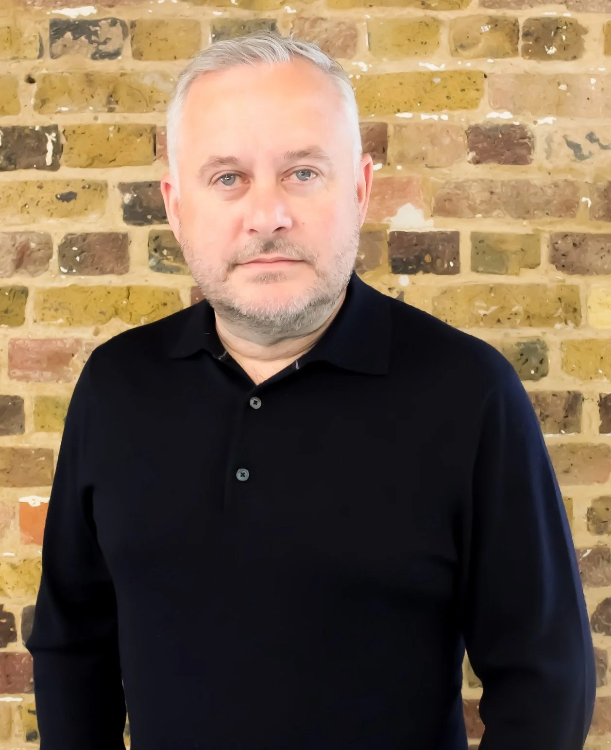 Portrait of a middle-aged man with light hair and beard standing in front of a brick wall.