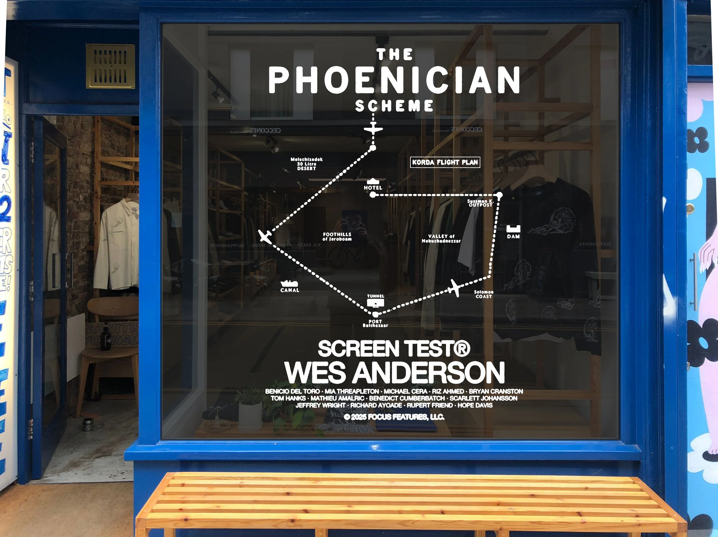 Store window display with a graphic map titled 'The Phoenician Scheme' showing locations like Gorden Flight Plan, Valley of Mebuchadnezzar, and Soloman Coast, and text indicating an art piece titled 'Screen Test' by Wes Anderson. The window has a blu
