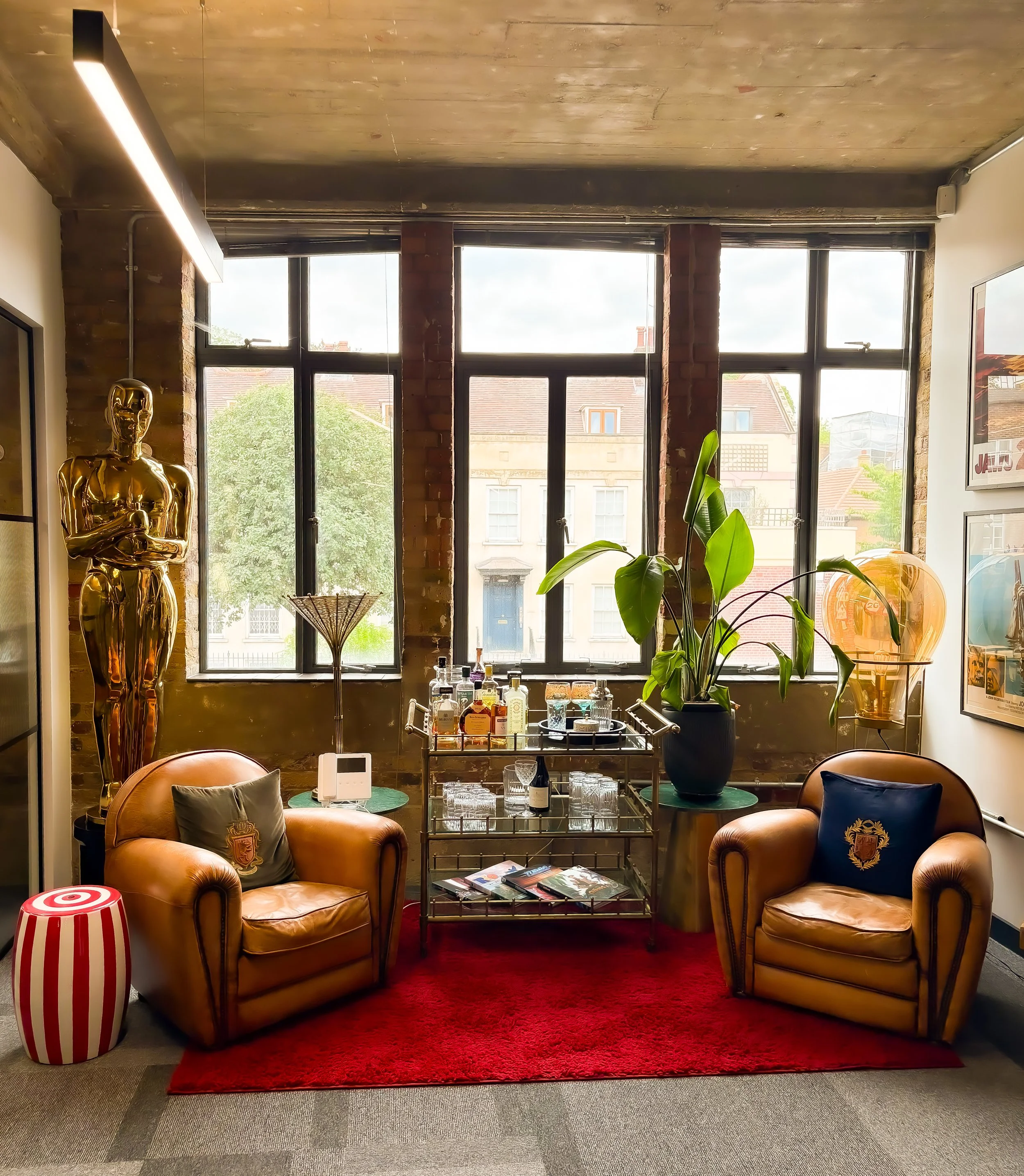 Contemporary lounge area with two tan leather armchairs with pillows, a red rug, a bar cart with bottles and glasses, a large potted plant, a gold sculpture of an Oscar-like figure, a striped side table, and large windows with brick and glass panes.