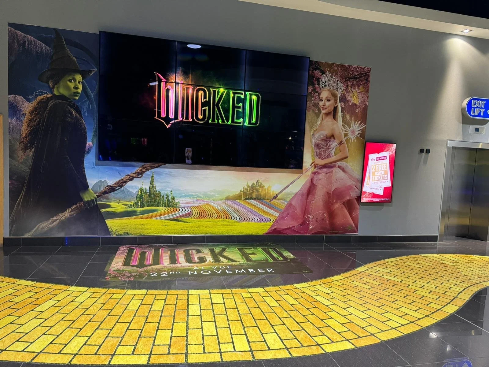 Advertisement for the musical 'Wicked' featuring characters from the show, with a colorful landscape background and promotional details on a digital screen.