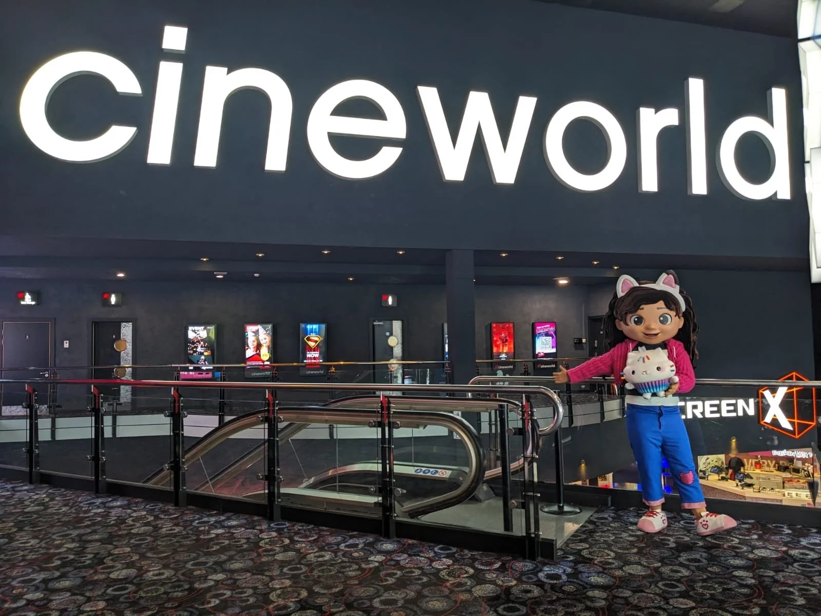Interior of a cinema with a large illuminated 'cineworld' sign on the wall. A cartoon doll with dark hair, a pink jacket, blue pants, and pink shoes is standing on the right, holding a white and pink cupcake plush toy. An escalator is visible in the
