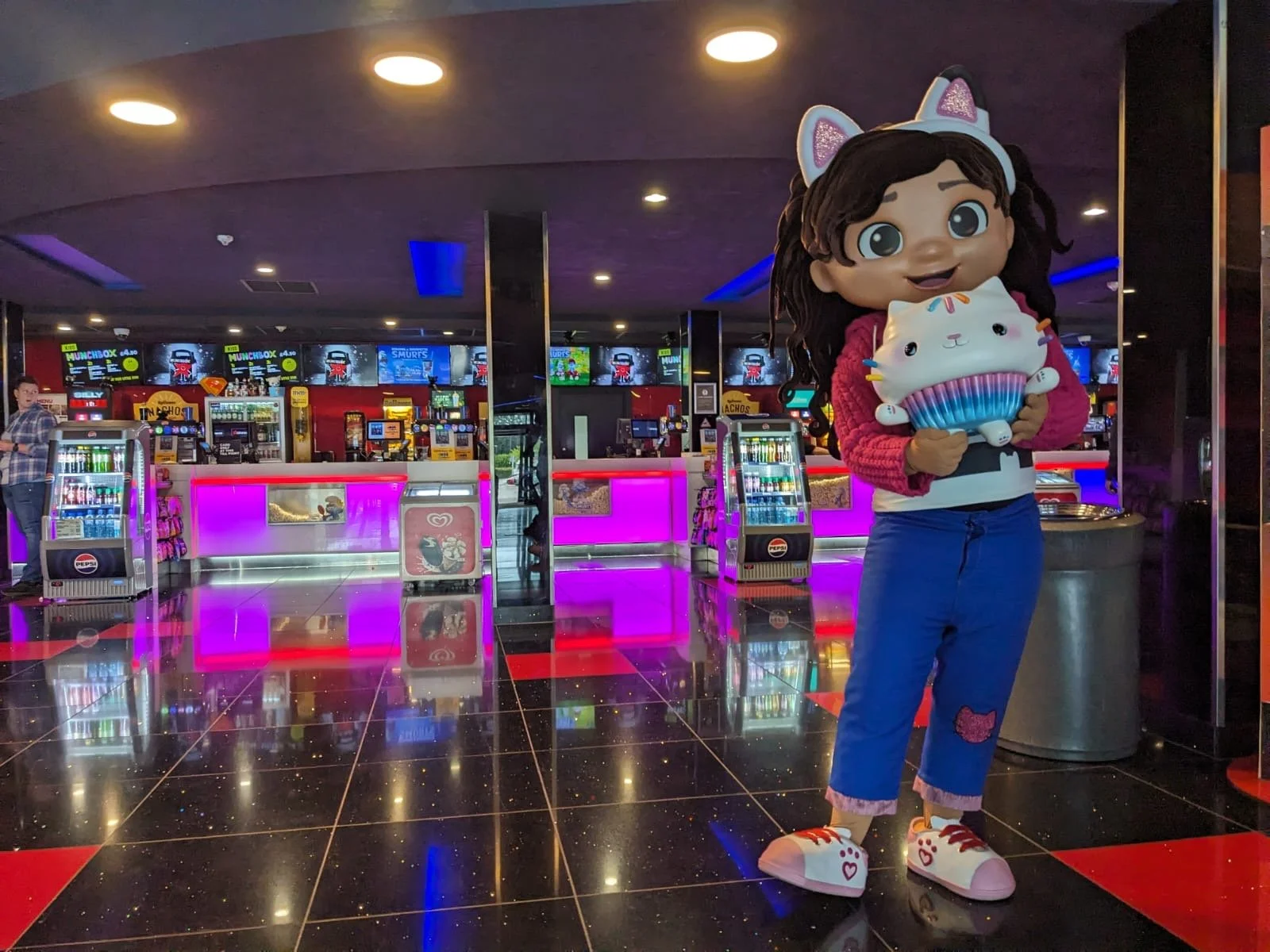 A person in a character costume of a girl with brown hair, blue eyes, wearing white cat ears, a pink sweater, blue jeans with a pink patch, and white sneakers with pink hearts, is standing in an arcade or entertainment center holding a large plush cu