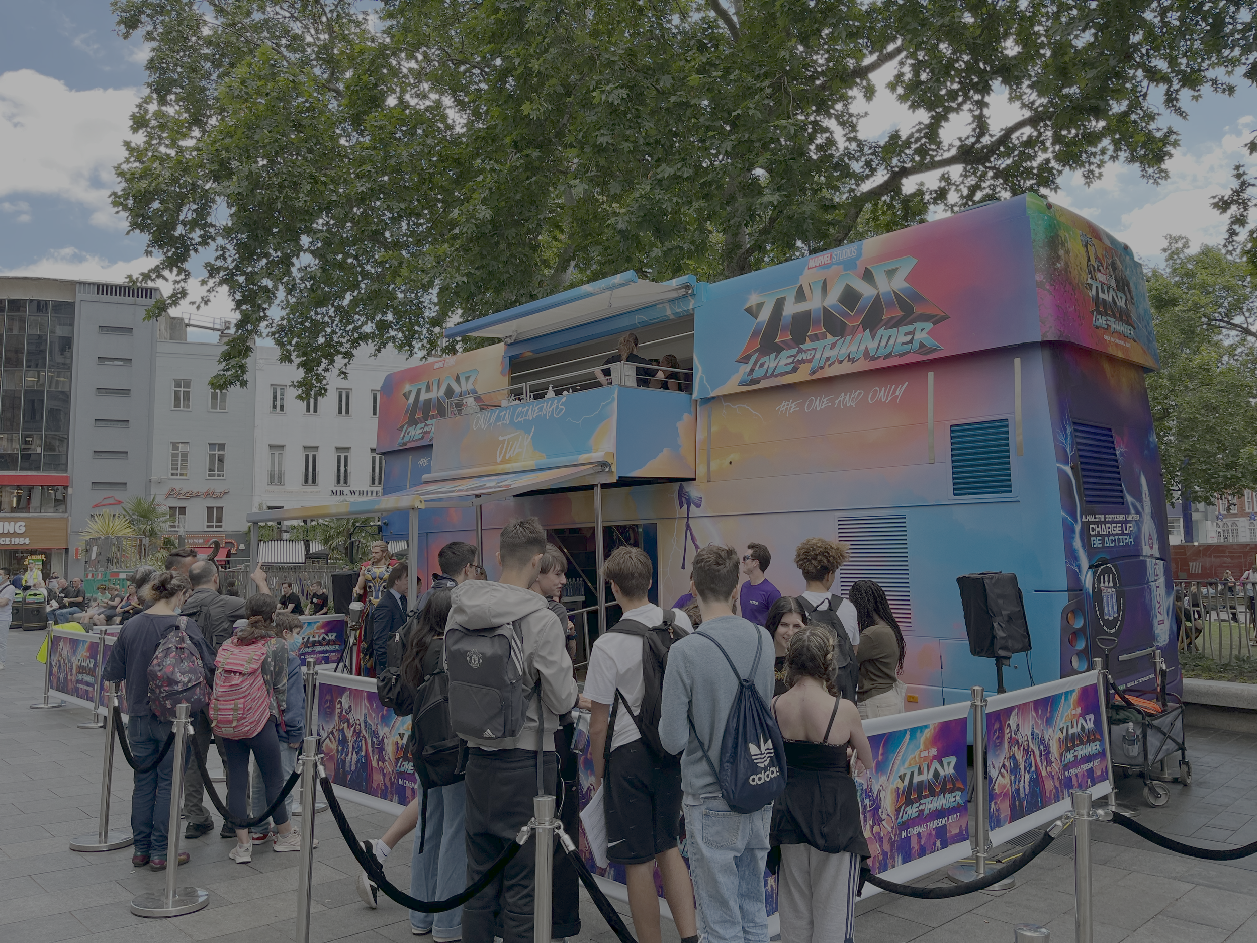 A line of people waiting outside a colorful Thor: Love and Thunder promotional bus in an urban setting, with trees and buildings in the background.