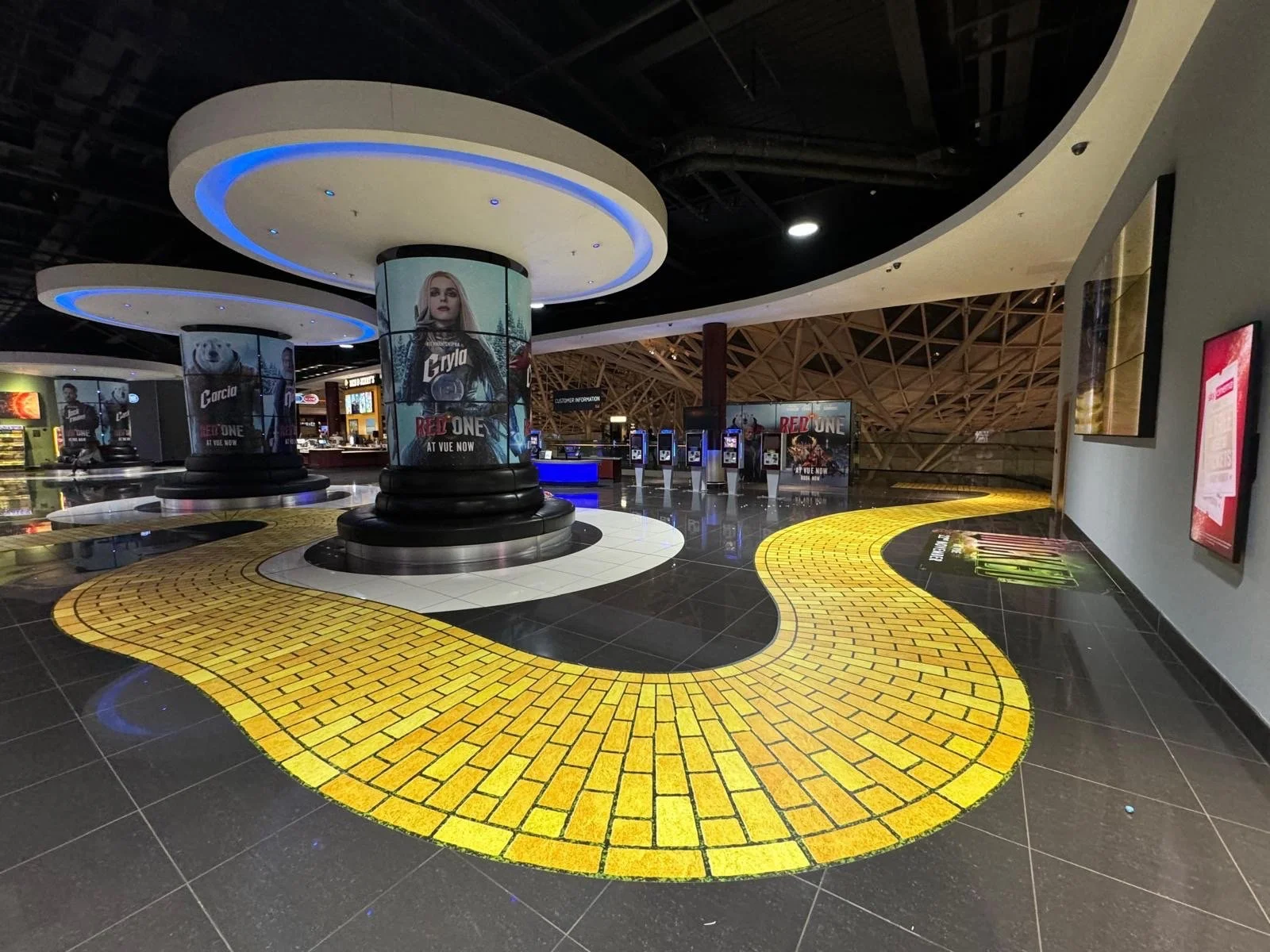 Empty airport terminal with a yellow brick-patterned pathway, digital advertisement screens, and a customer service area.