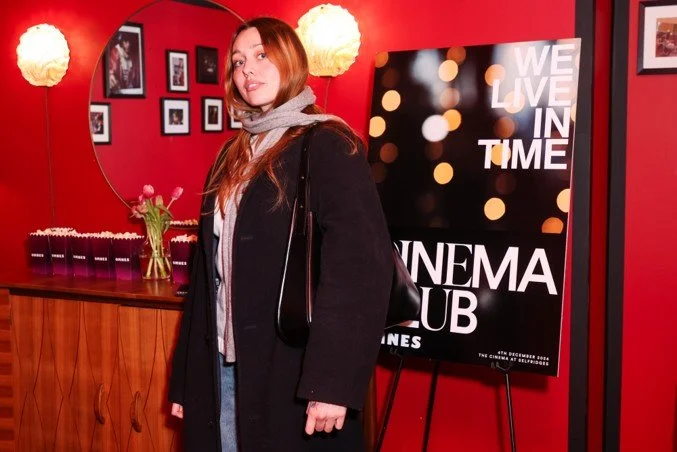 Young woman standing in cinema club decorated with red walls, framed photos, and floral centerpiece, near a sign that reads 'We Live in Time Cinema Club'.