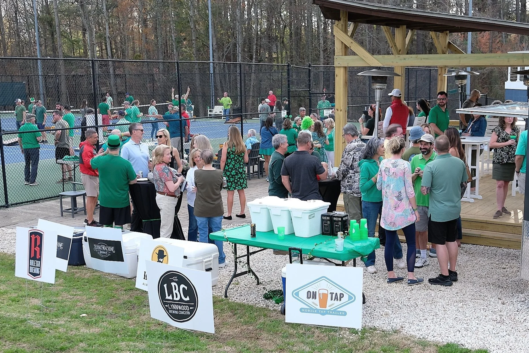 People socializing outside near a baseball field with a chain-link fence, some on a deck area, and others playing or watching a game. There are signs for local breweries and a mobile tap trailer.