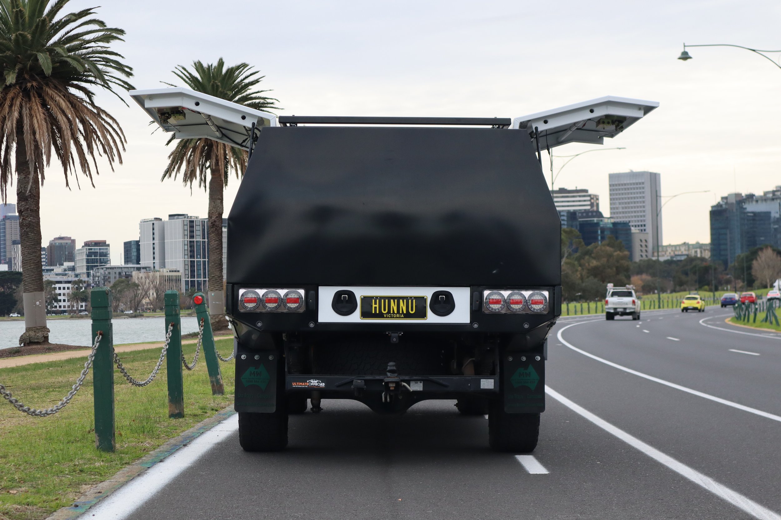 Black utility vehicle parked on a city street near a park with palm trees, with city buildings in the background.