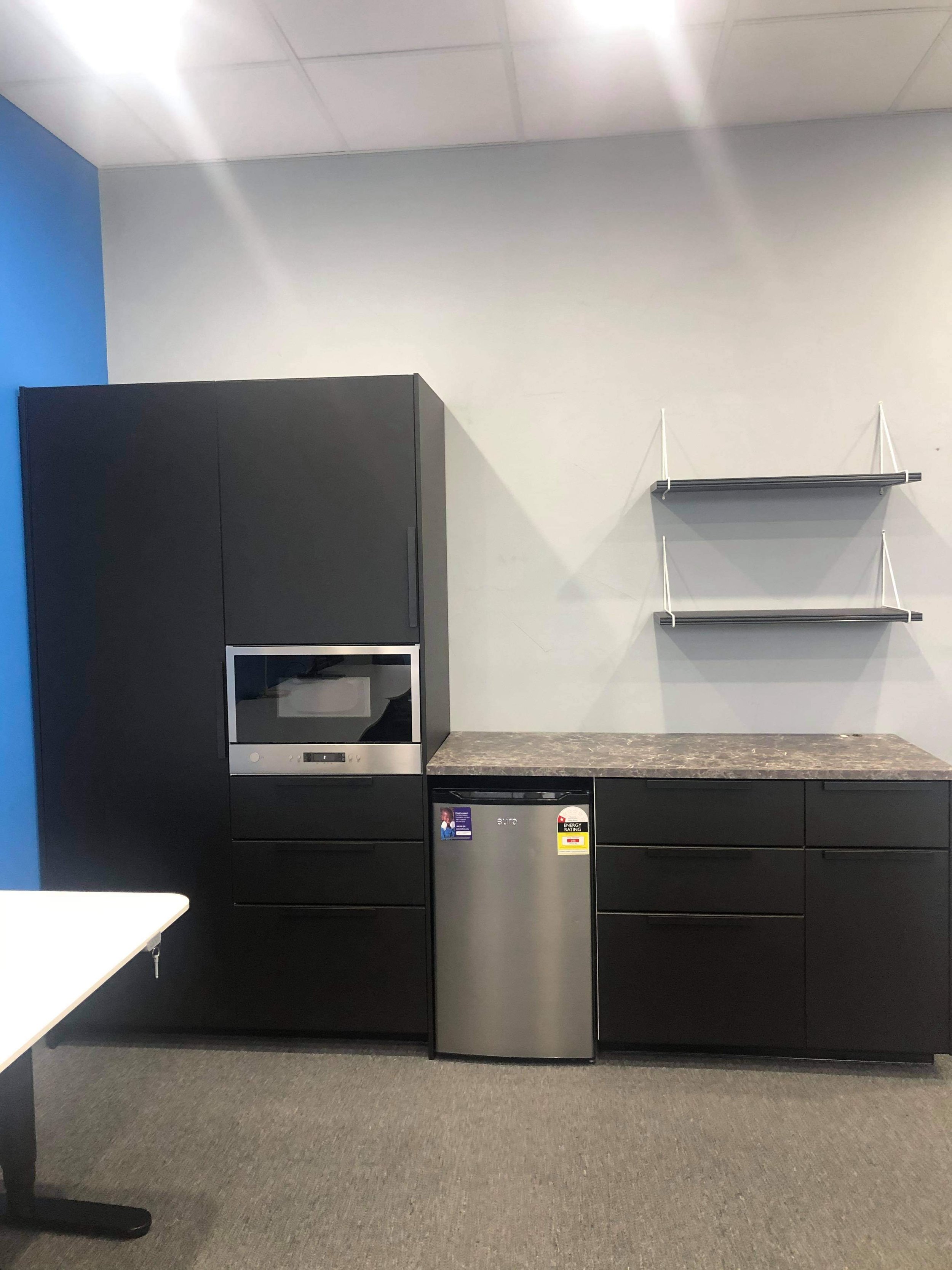 Black kitchen cabinets with a microwave, a small mini refrigerator, and open shelves on a white wall, with a blue accent wall on the left. There is a gray countertop and a gray carpeted floor.