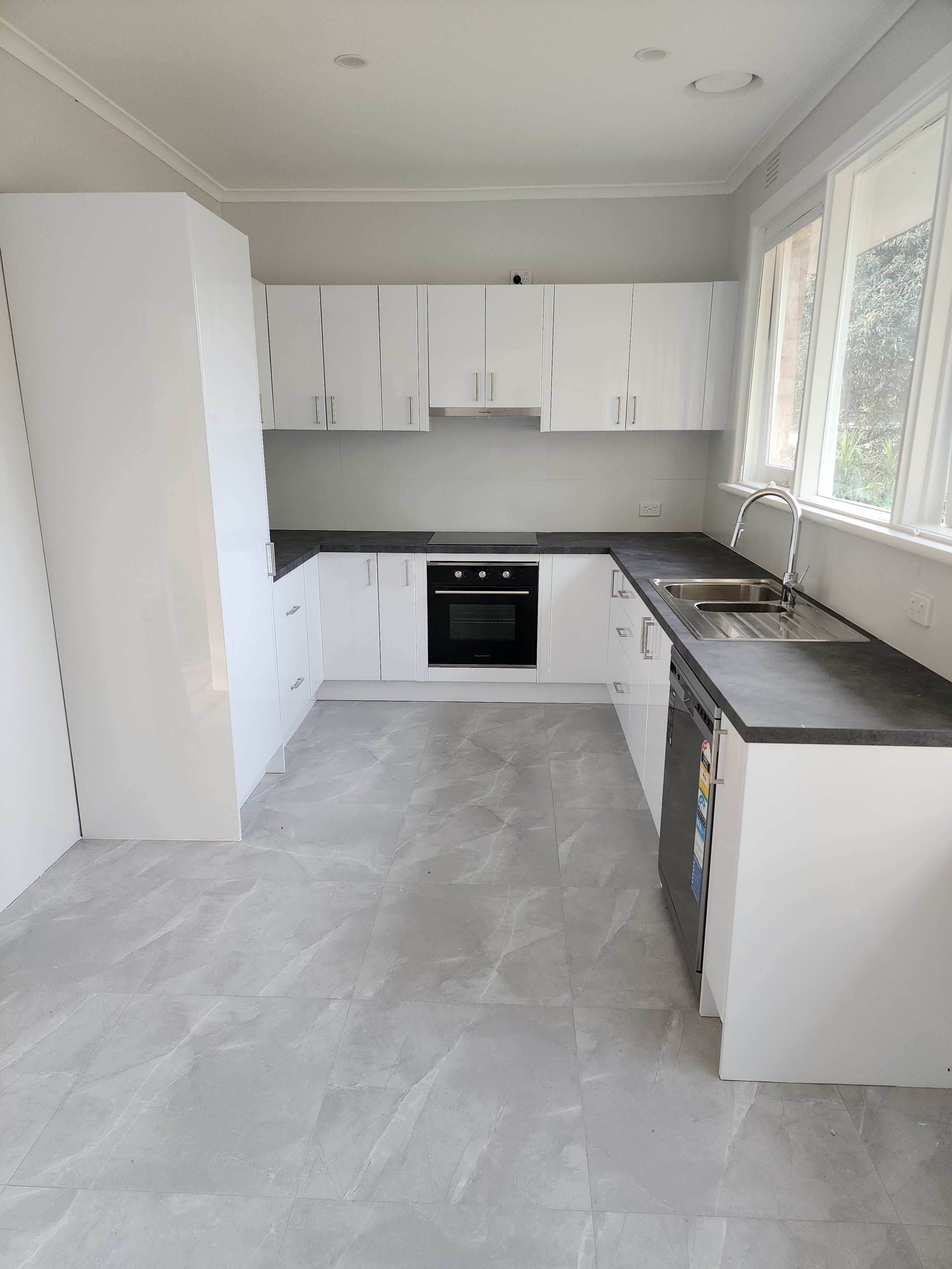 Empty modern kitchen with white cabinets, black countertop, gray tiled floor, stainless steel sink, and large windows.