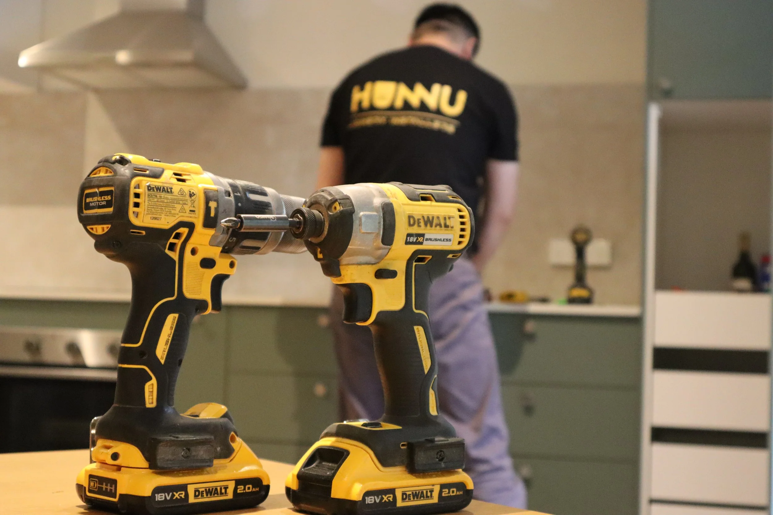 Two yellow and black DEWALT cordless power tools on a wooden surface with a person working in a kitchen background.