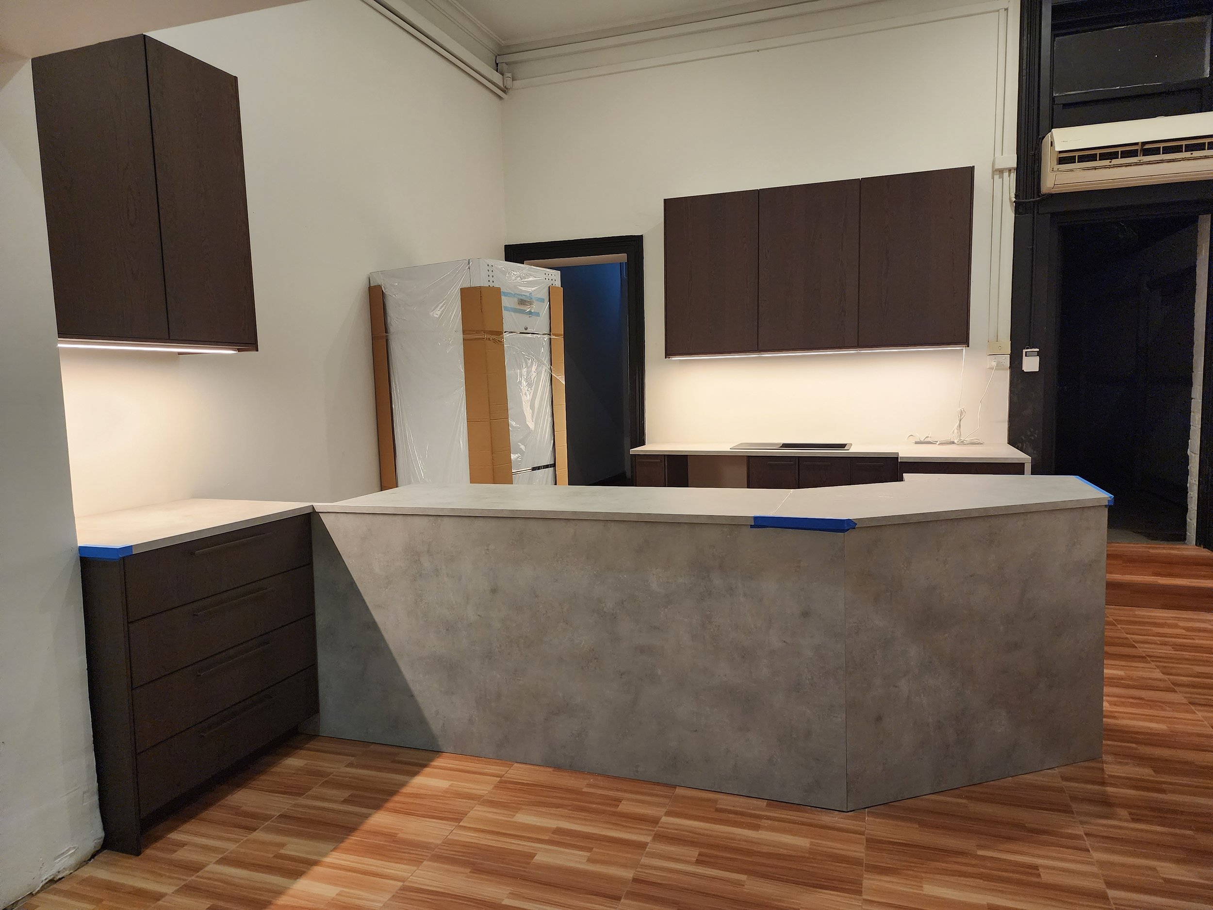 Kitchen area with dark brown cabinets, a concrete counter, wooden flooring, and a refrigerator wrapped in plastic, possibly under construction or renovation.
